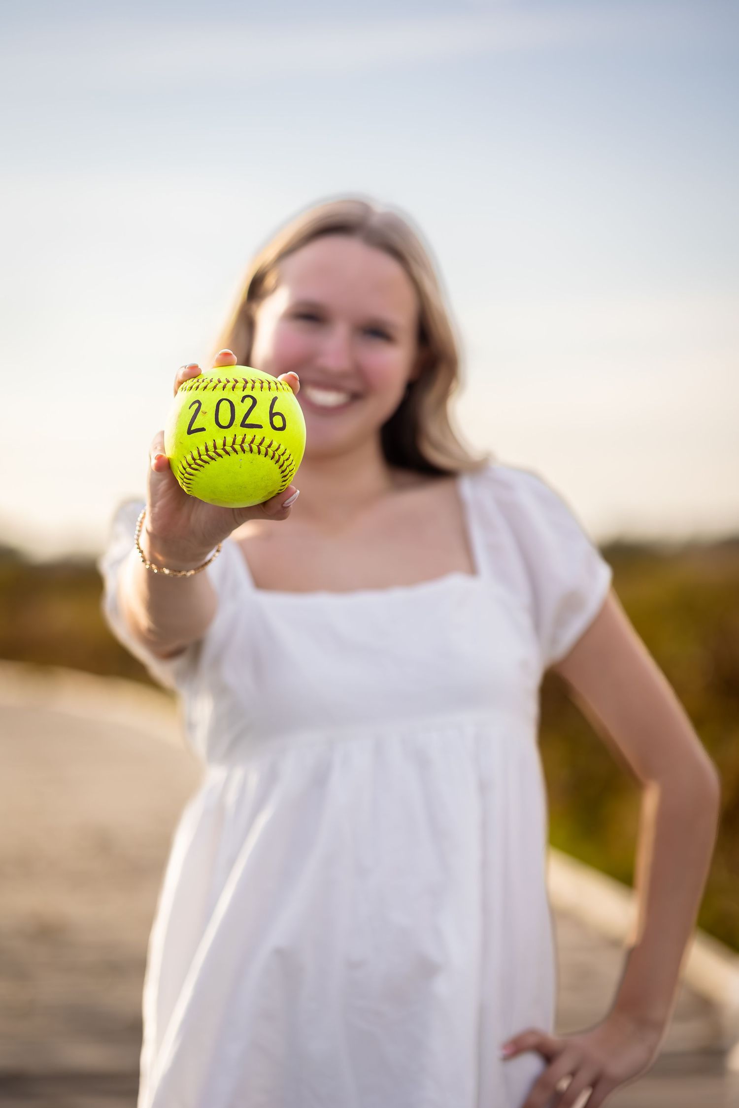 A person in a white dress holds up a yellow softball with '2028' written on it against a sunset background.