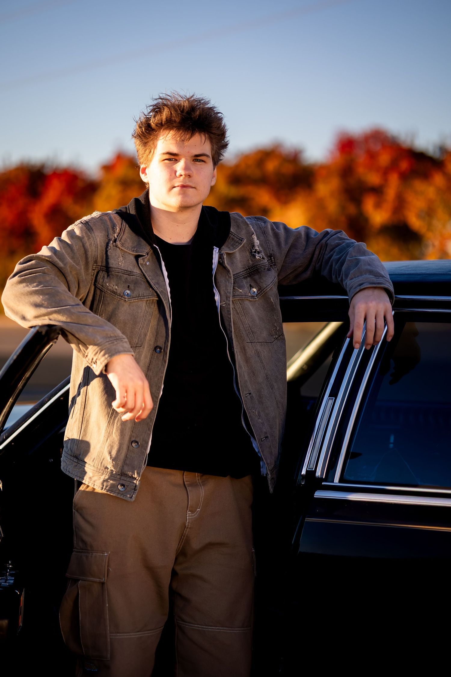 Casual autumn photoshoot featuring someone leaning against a black car with fall foliage in the background.