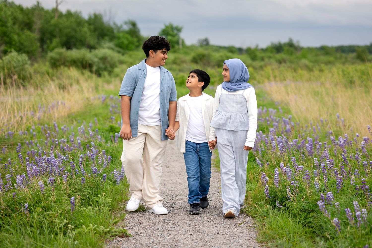 A Muslim family of three walks hand-in-hand on a path through a field of purple wildflowers.