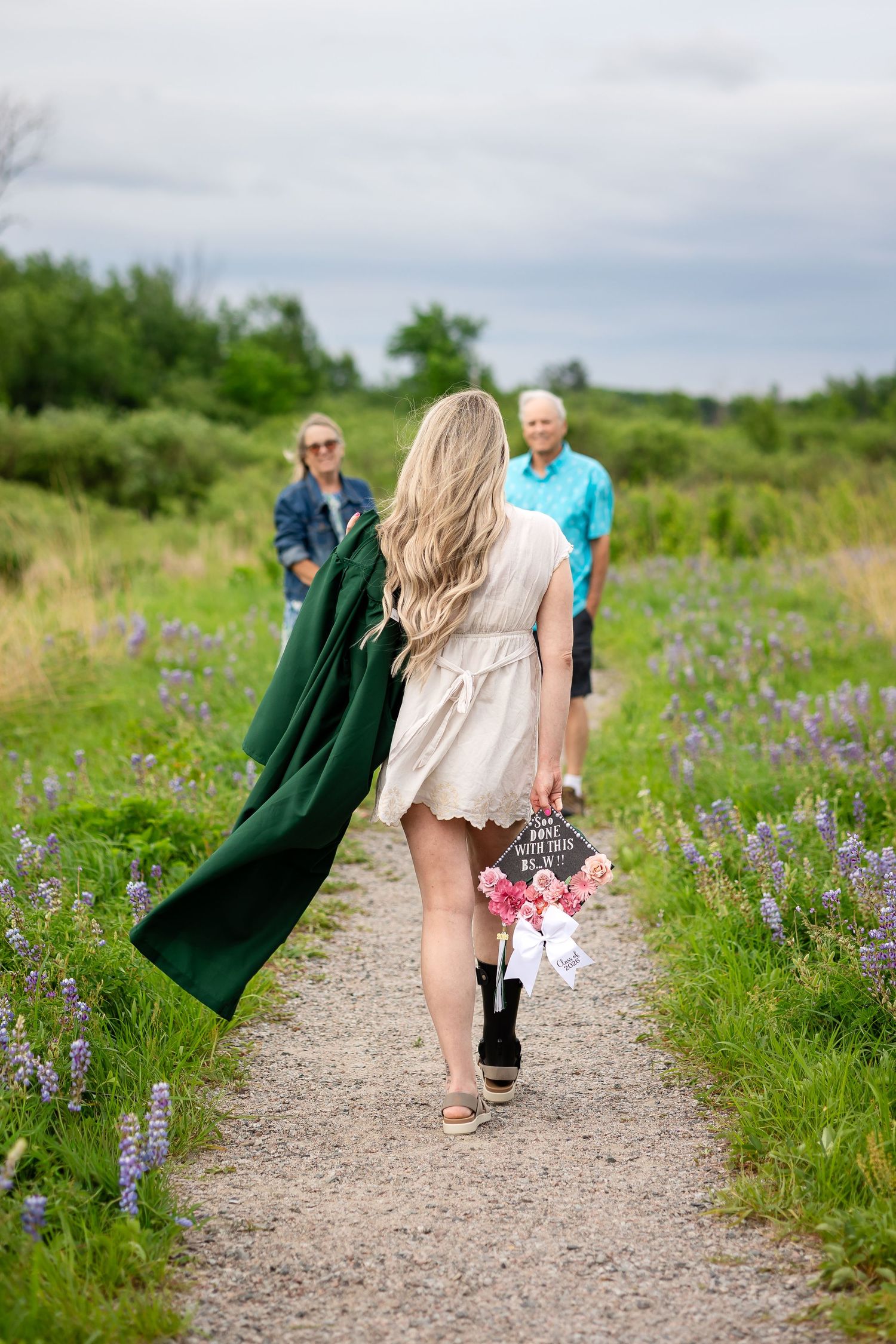Graduate in green cap and gown walks along a scenic wildflower path toward smiling grandparents in a lush field.