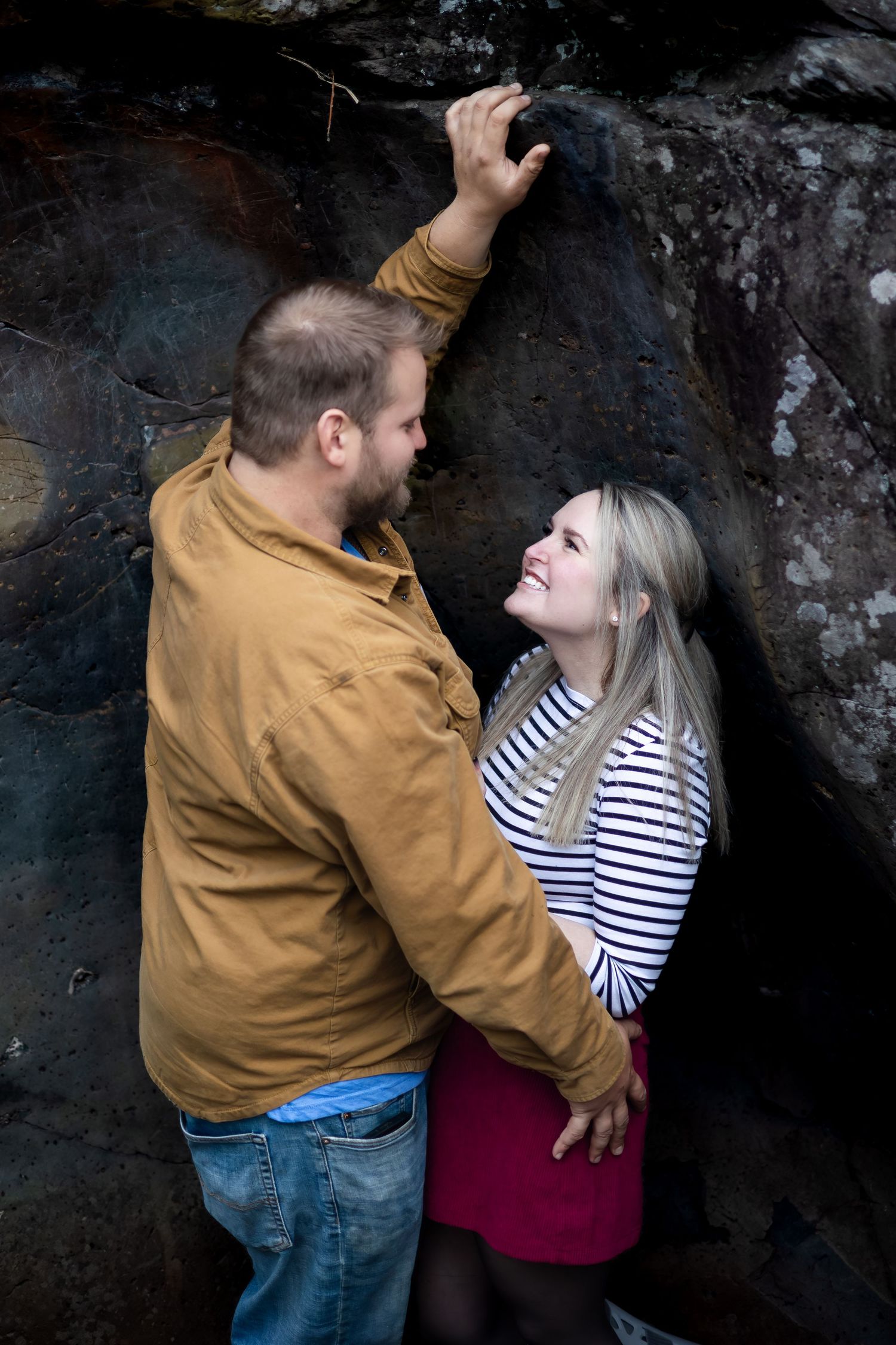 Couple sharing a romantic moment against a rocky backdrop, man in brown jacket leans toward smiling blonde woman in striped top.