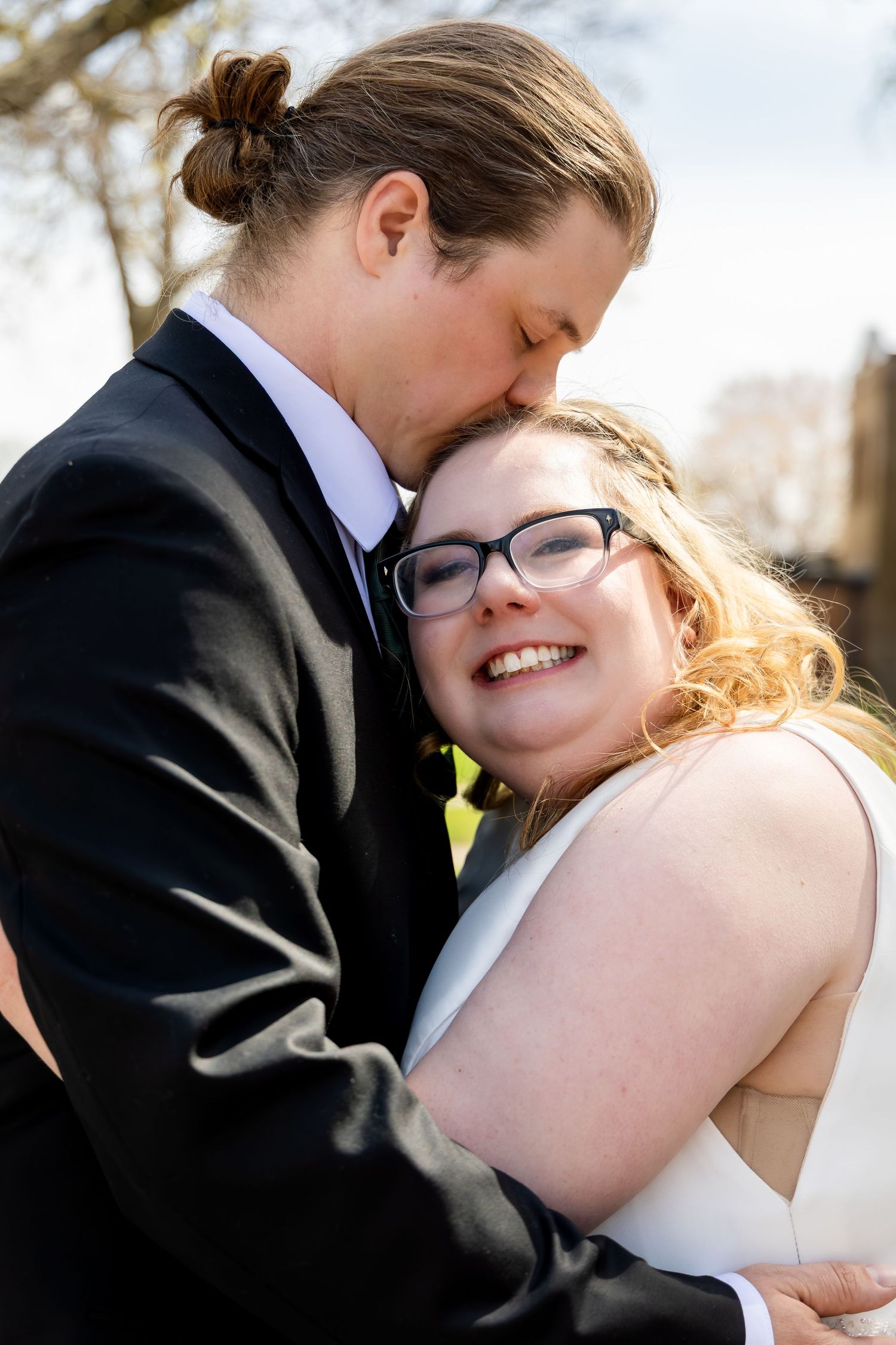 Groom in black suit kissing smiling bride with glasses on her cheek during outdoor wedding portraits.