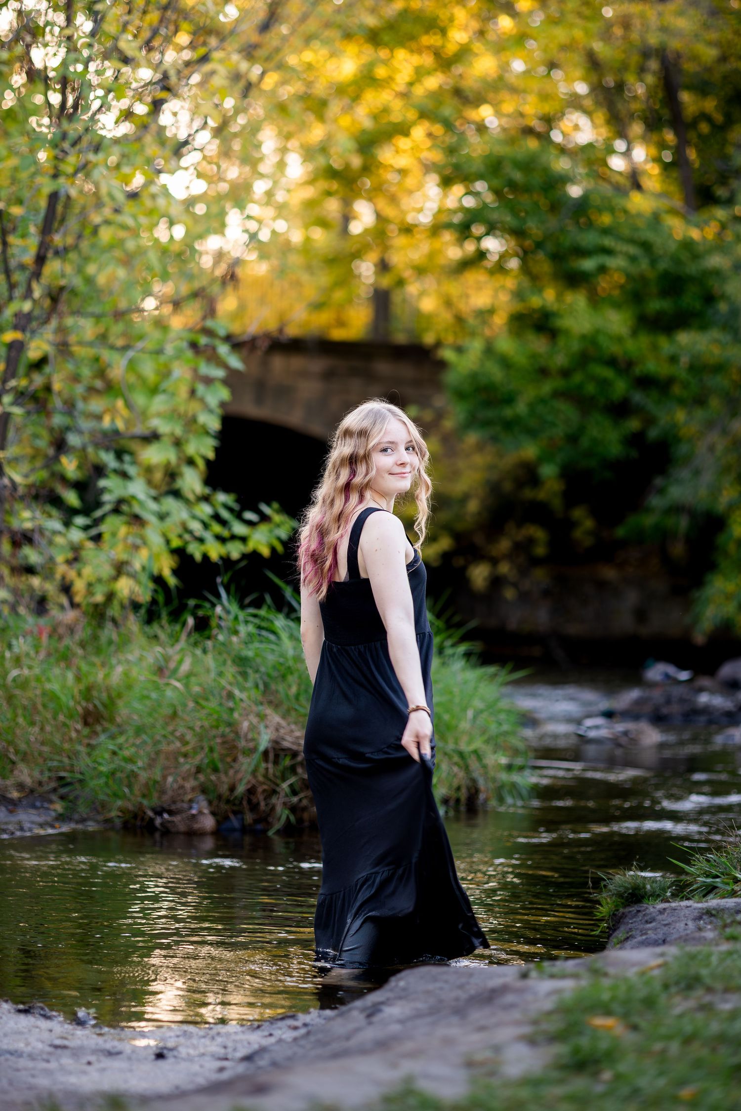 A long black dress flows in a shallow creek surrounded by autumn foliage and a stone bridge in the background.