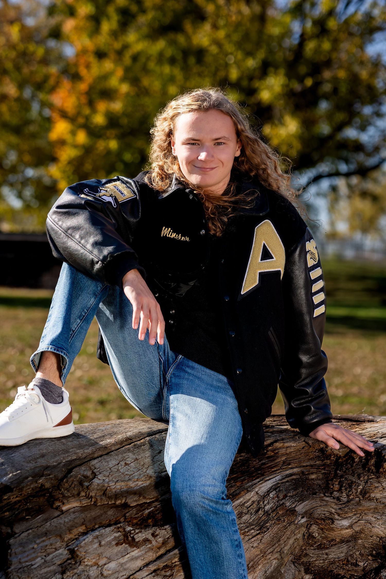 Person in black jacket and jeans poses casually on fallen tree trunk during autumn day with golden foliage in background.