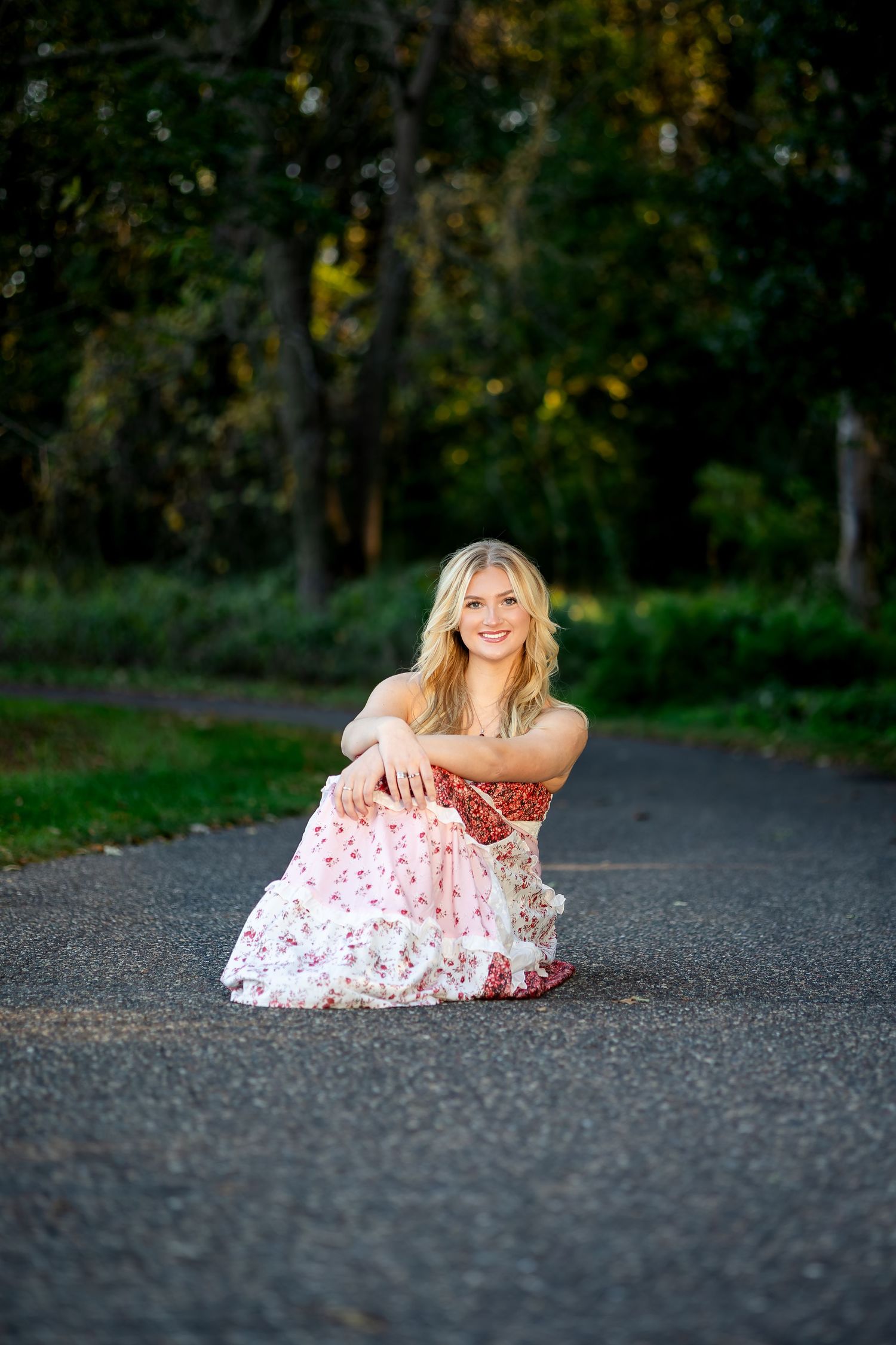 A person in a floral dress sits on a paved path surrounded by lush greenery at sunset.