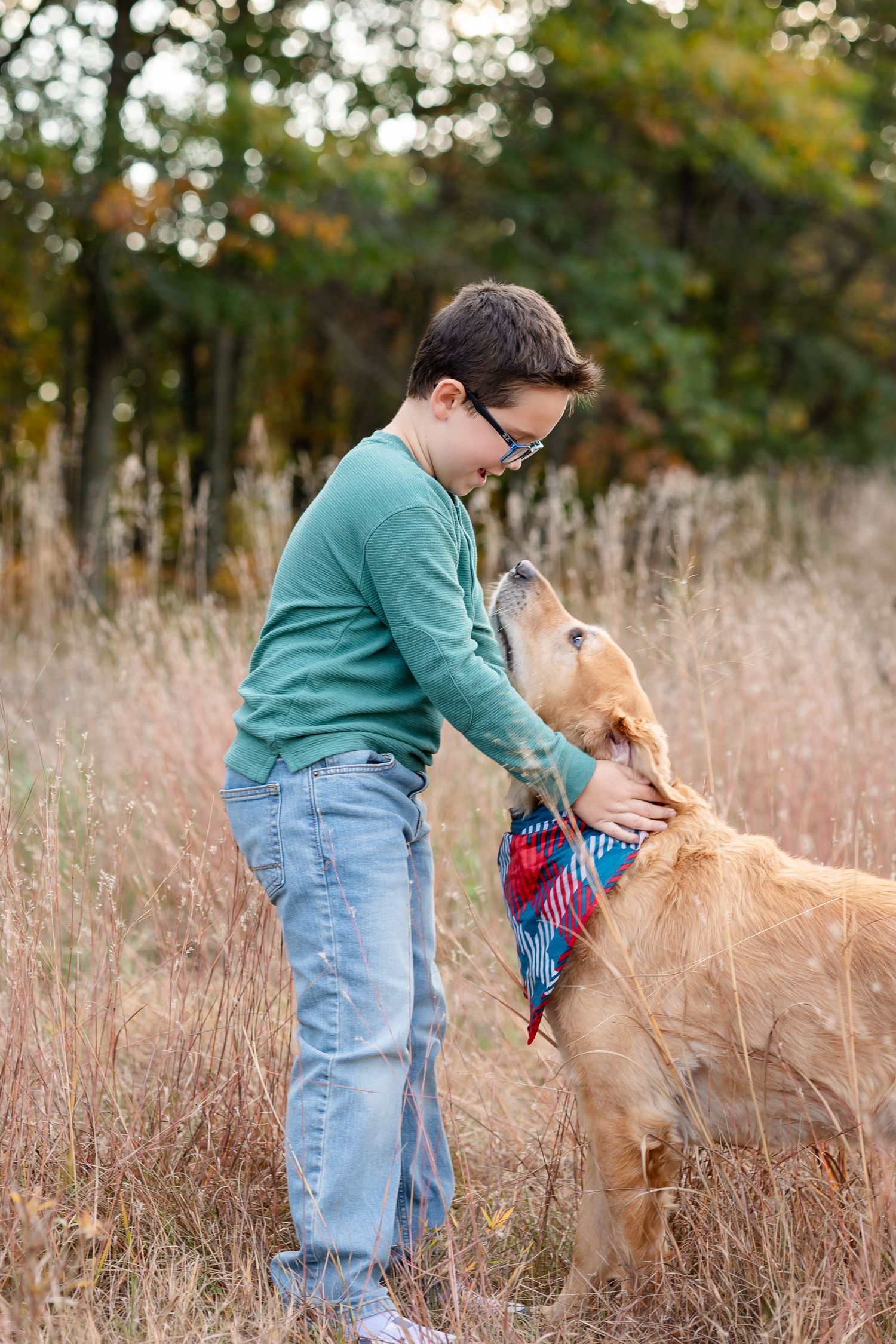 A person in a teal sweater pets a golden retriever wearing a bandana in a field of tall grass.