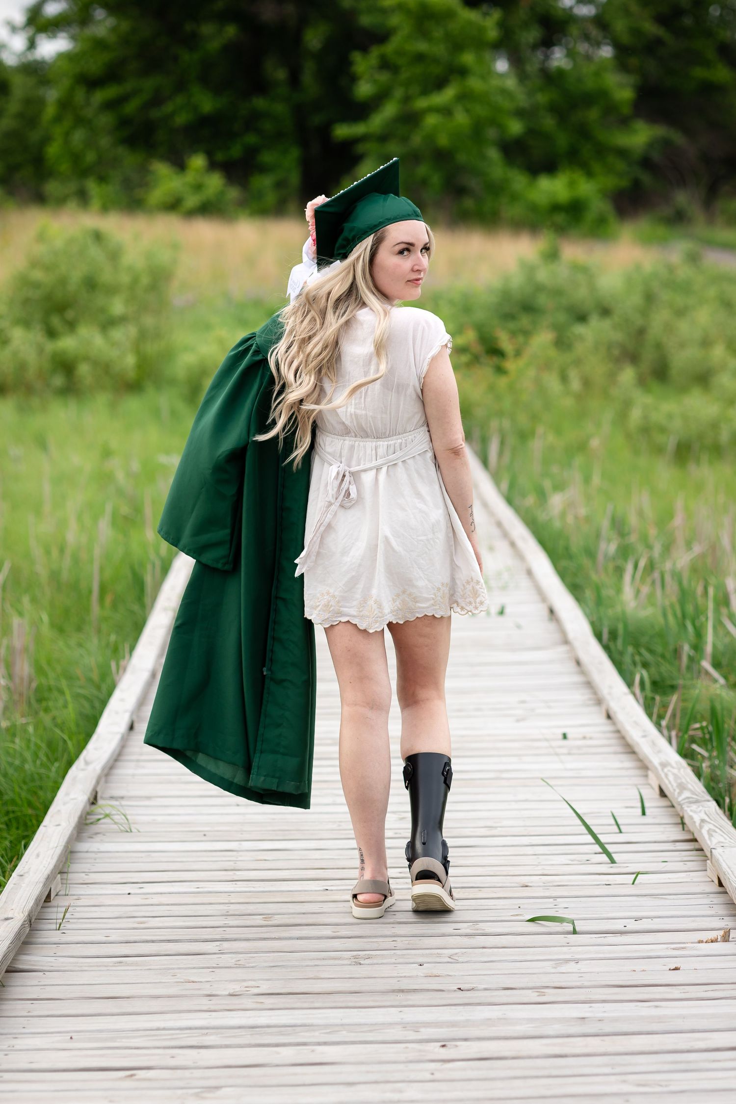 Blonde woman in white dress and green graduation cap and gown walks along wooden boardwalk path.