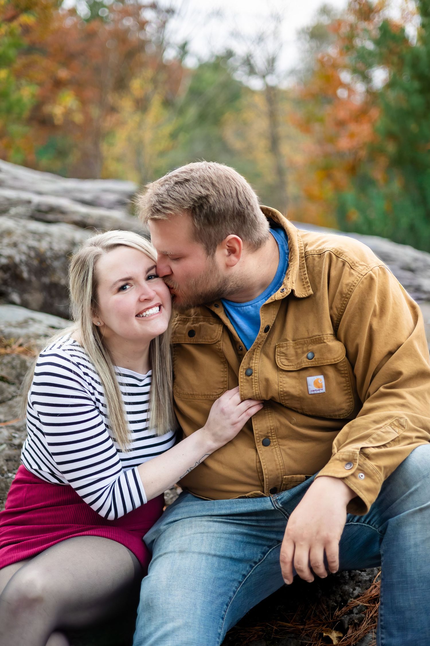 Man in brown jacket kissing smiling woman on cheek while sitting on rocks in fall foliage.