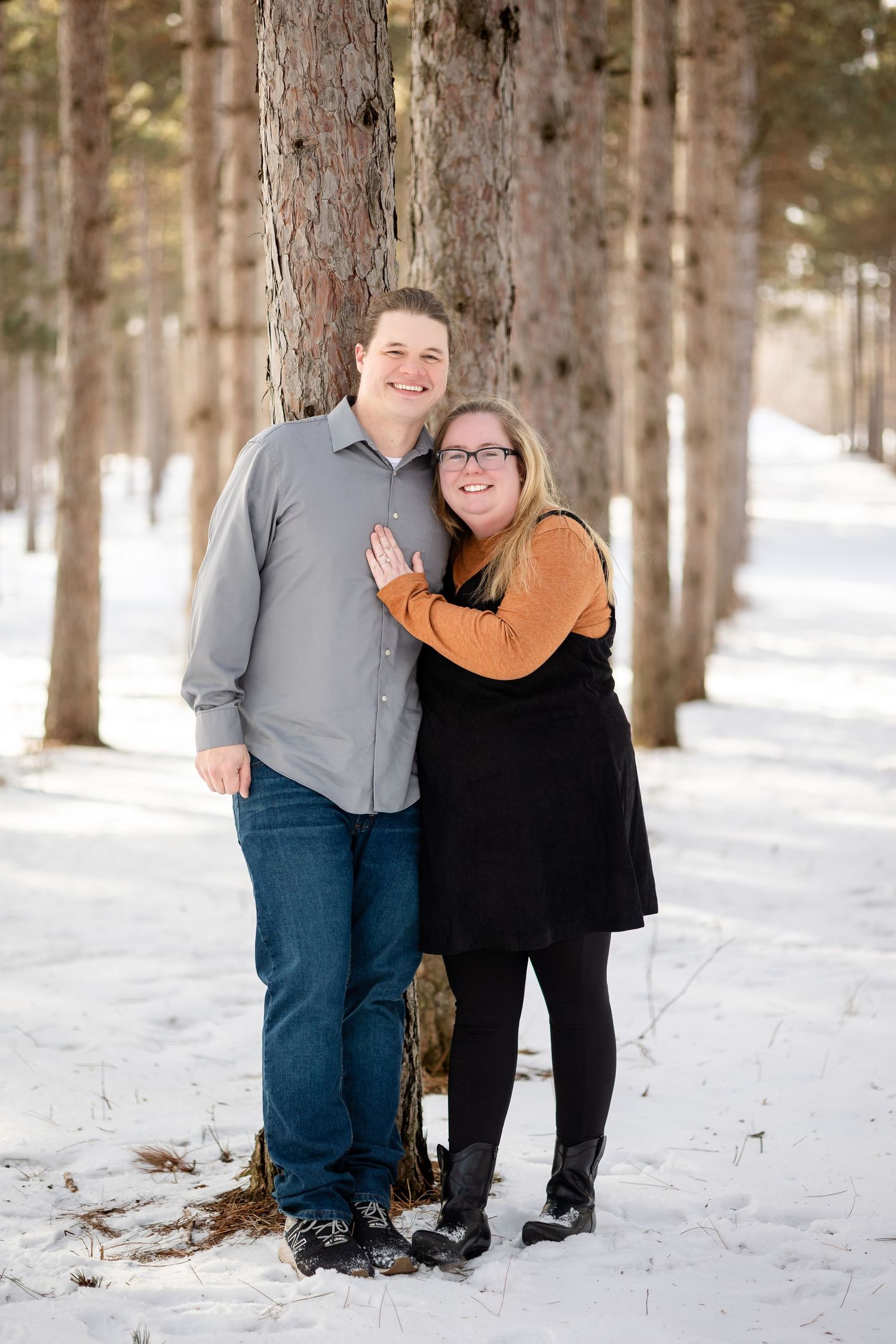 Engaged couple posing together against pine tree in snow-covered forest, woman showing engagement ring.