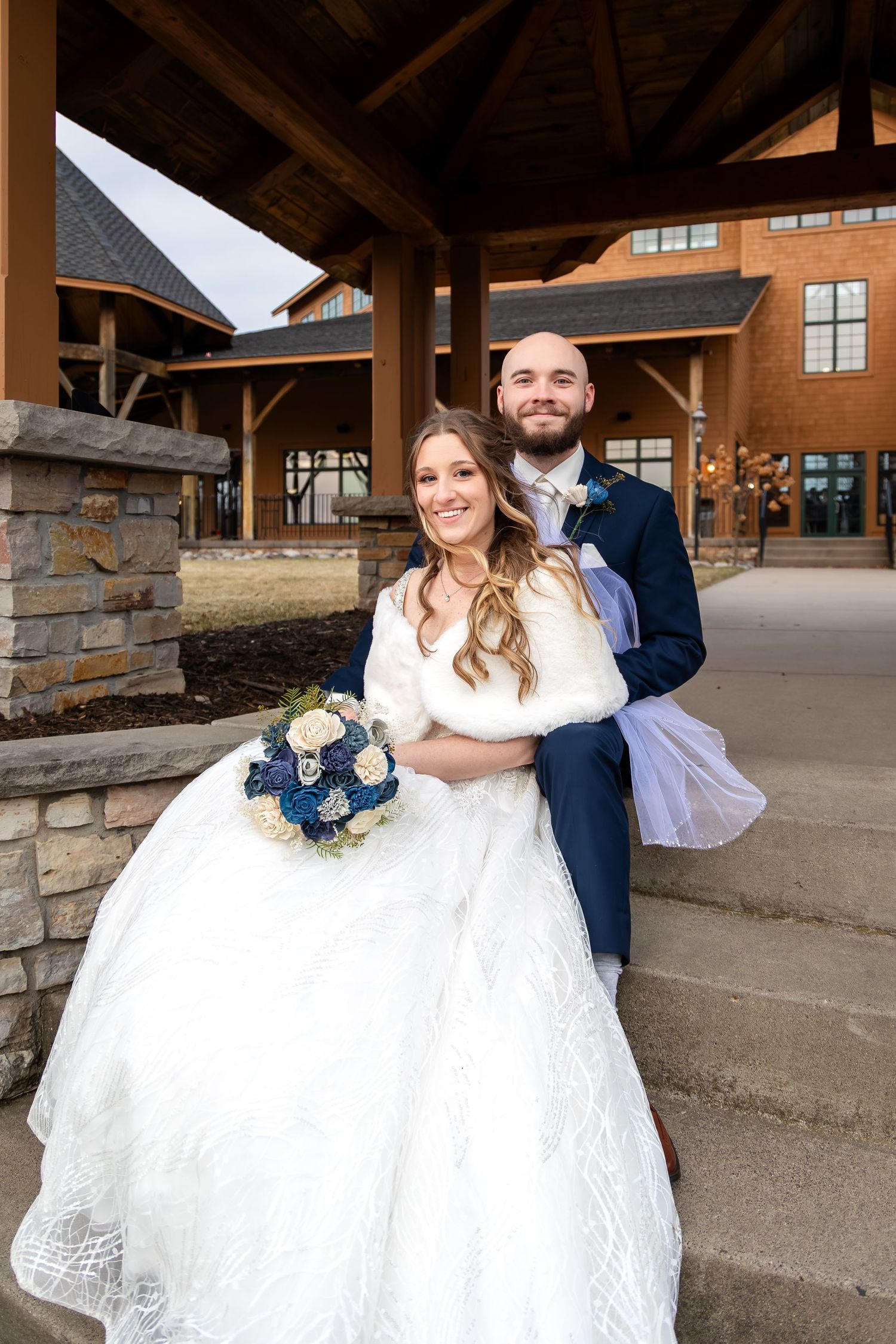 Bride in white gown with fur wrap holds blue bouquet as groom in navy suit embraces her outside rustic wedding venue.