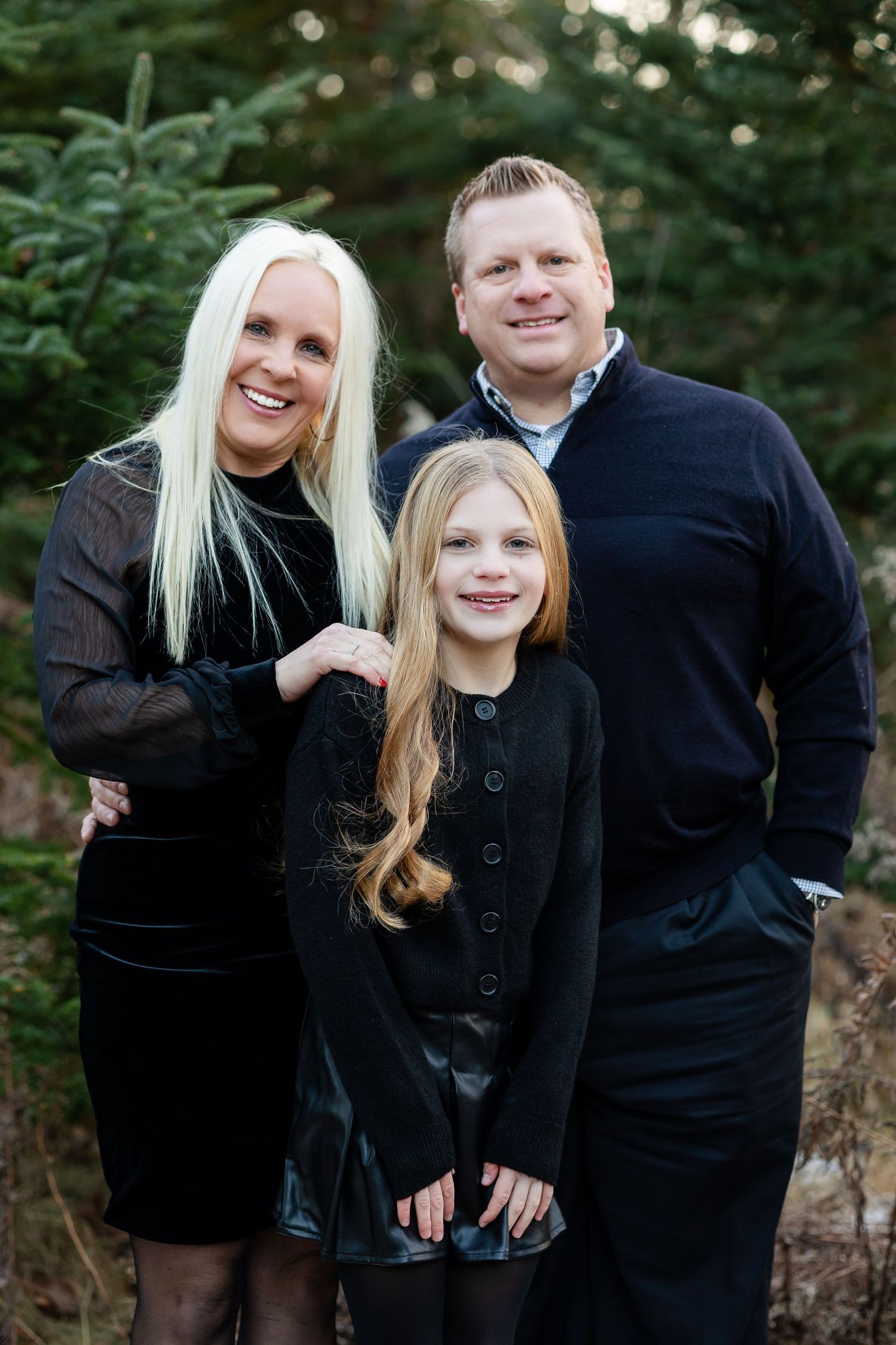 Family members wearing coordinated black outfits pose together against an evergreen forest backdrop.