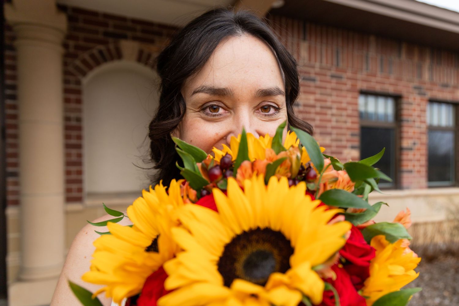 Bright sunflowers and red flowers create a colorful wedding bouquet against a brick backdrop.