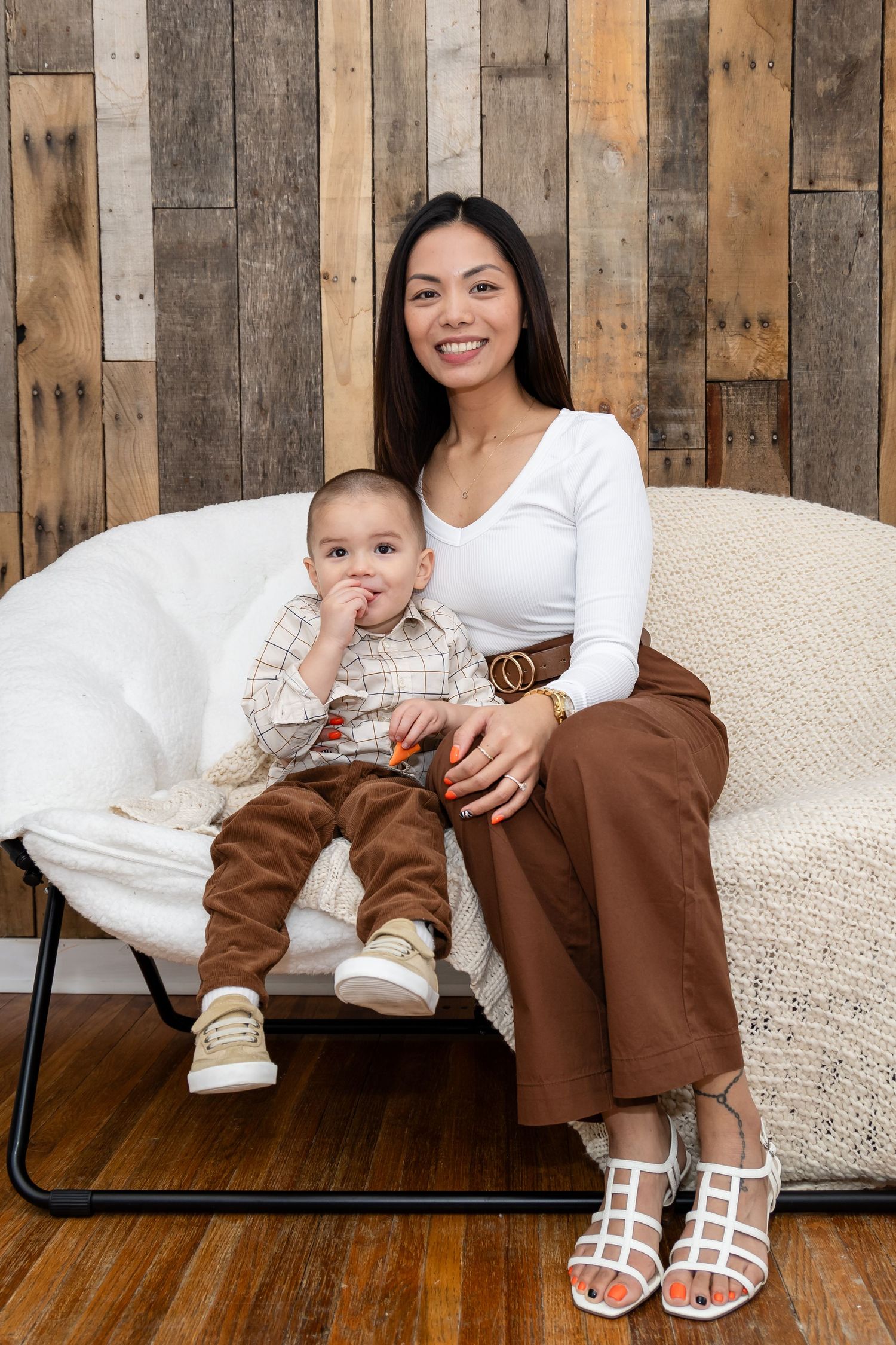 Family portraits on white couch against rustic wood panel wall wearing coordinated brown and white outfits.