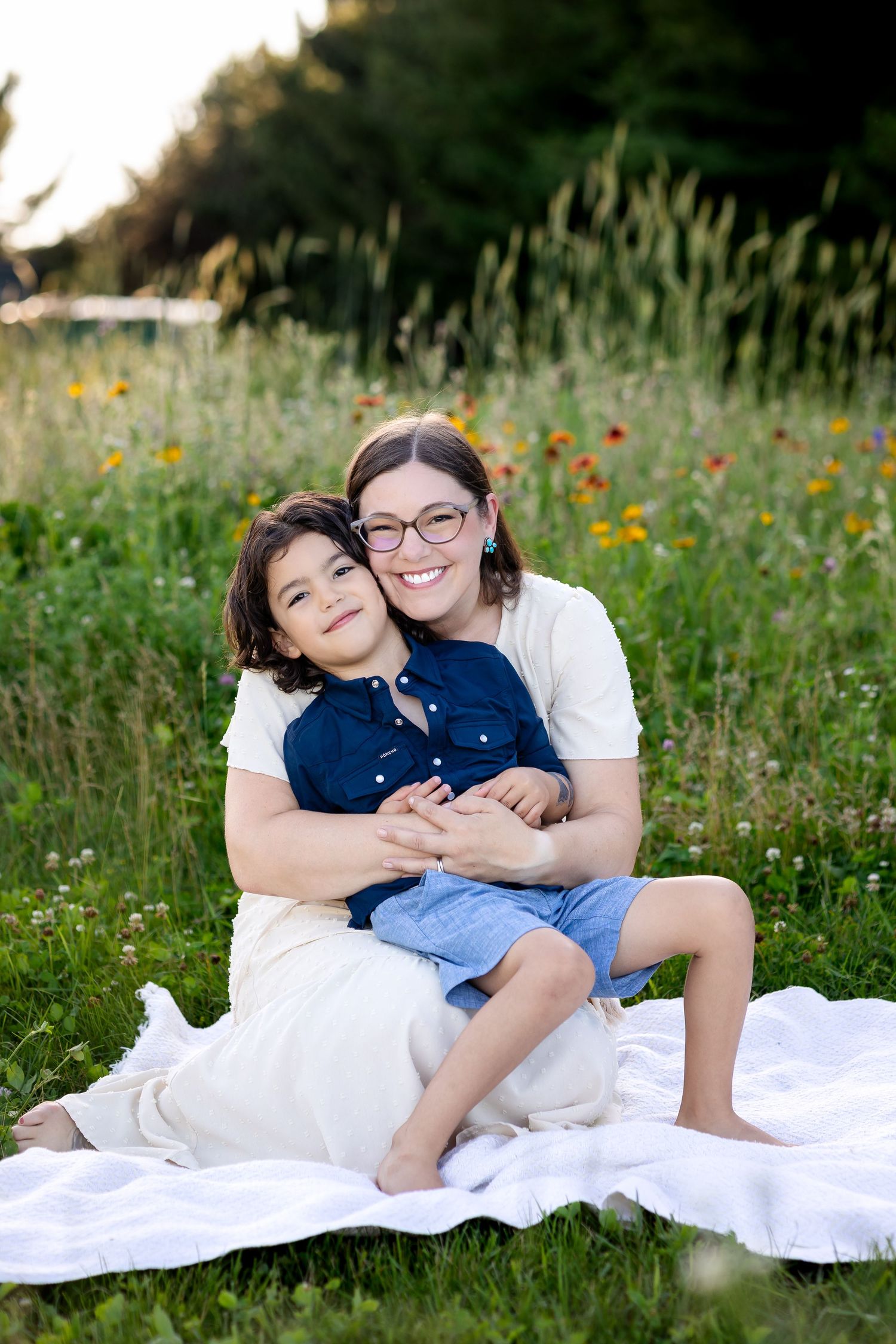 Two people share tender moments while sitting on a white blanket in a wildflower meadow at sunset.