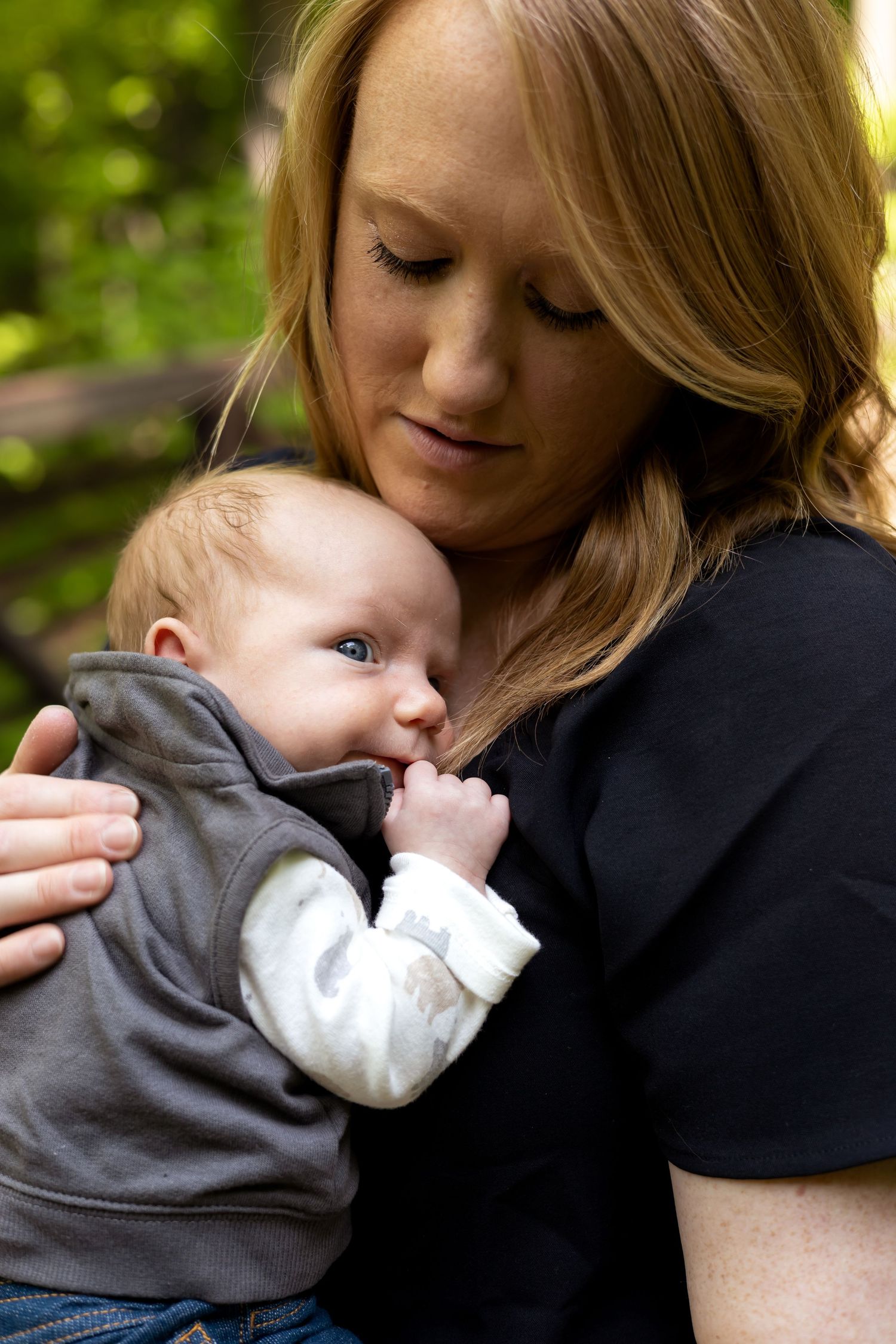 Tender moment between parent and infant in black and gray clothing against natural outdoor backdrop.