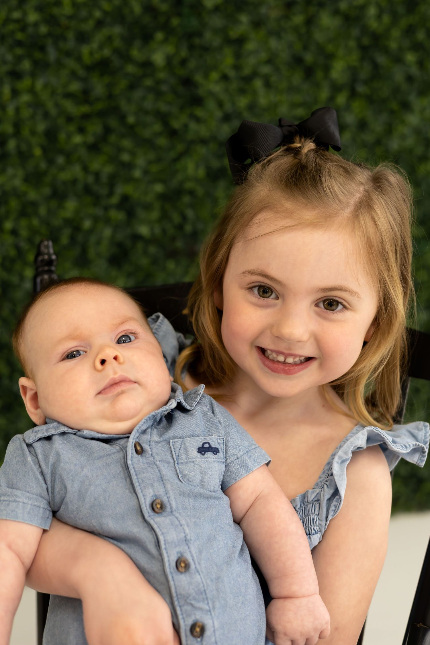 Two children in denim outfits posing together against a green leafy background.