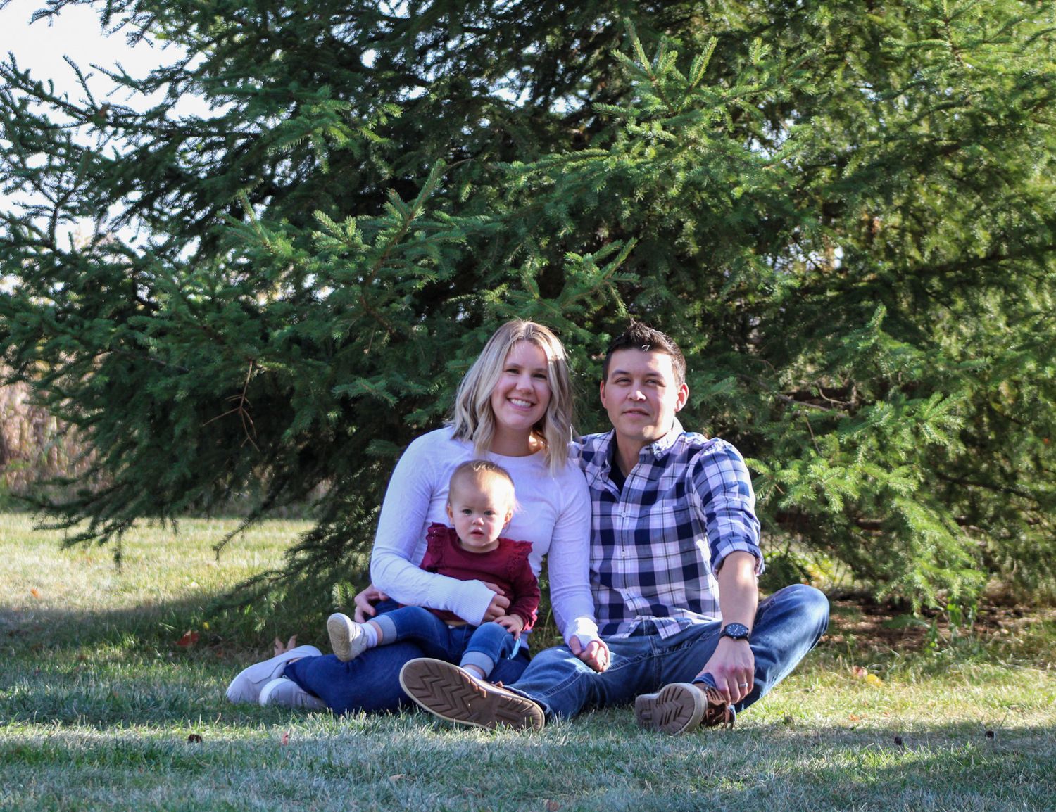 Family relaxing together on grass under evergreen trees during outdoor photo session.