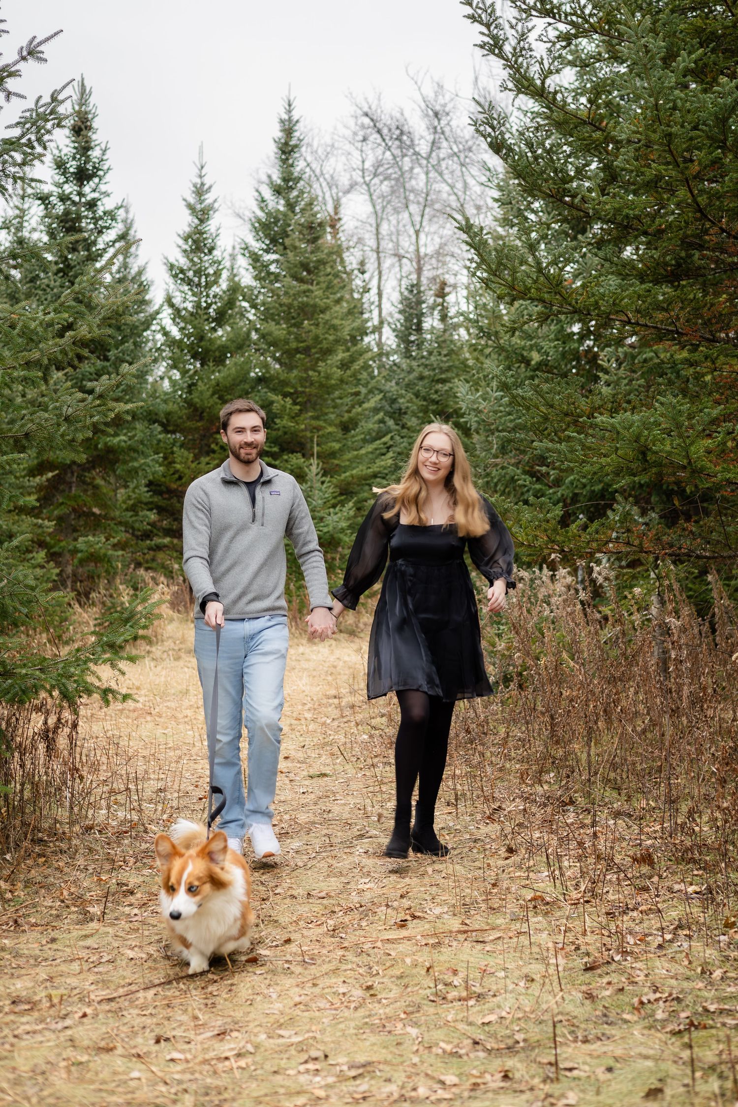 A couple walks their corgi dog along a trail surrounded by evergreen trees.