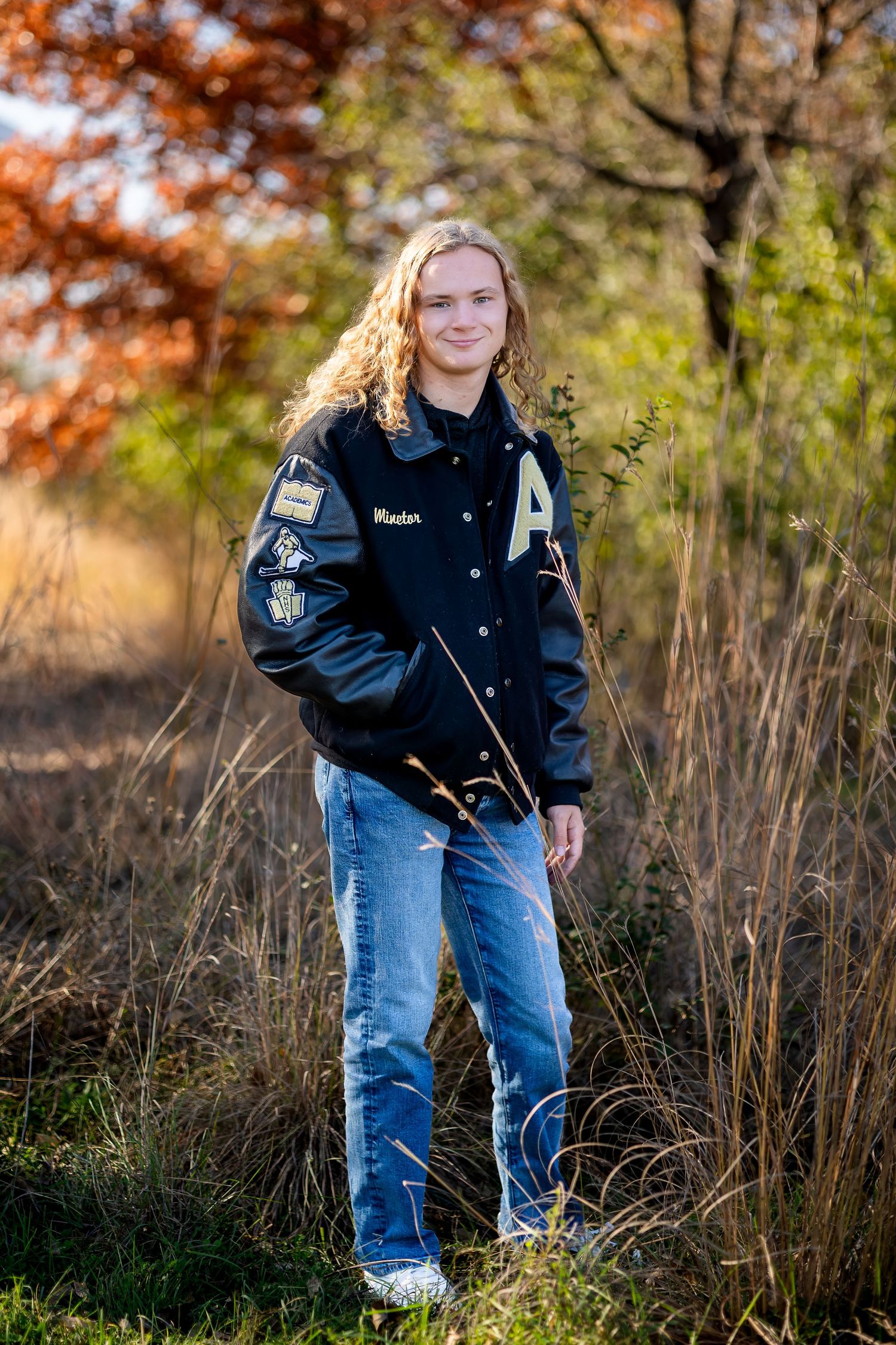 Student stands in tall grass wearing a varsity jacket against a backdrop of fall foliage and trees.