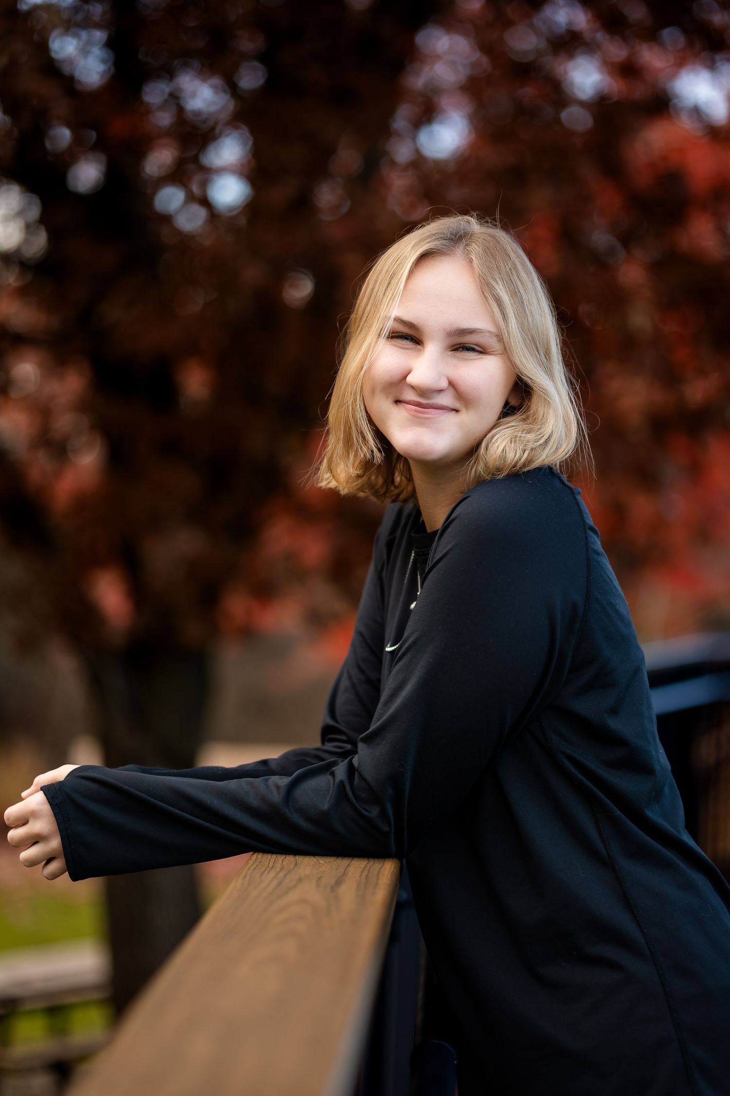 A person in a black sweater poses outdoors against a backdrop of autumn trees with warm orange foliage.