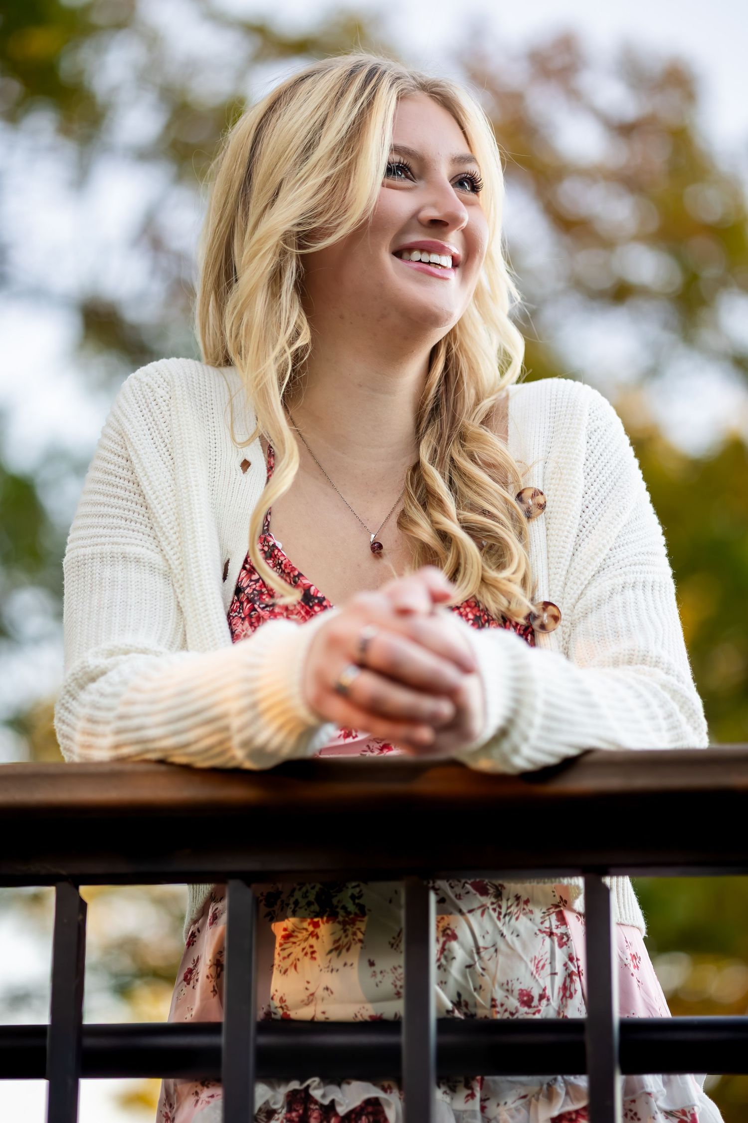 Woman in white sweater and floral dress leans on wooden railing during golden hour.