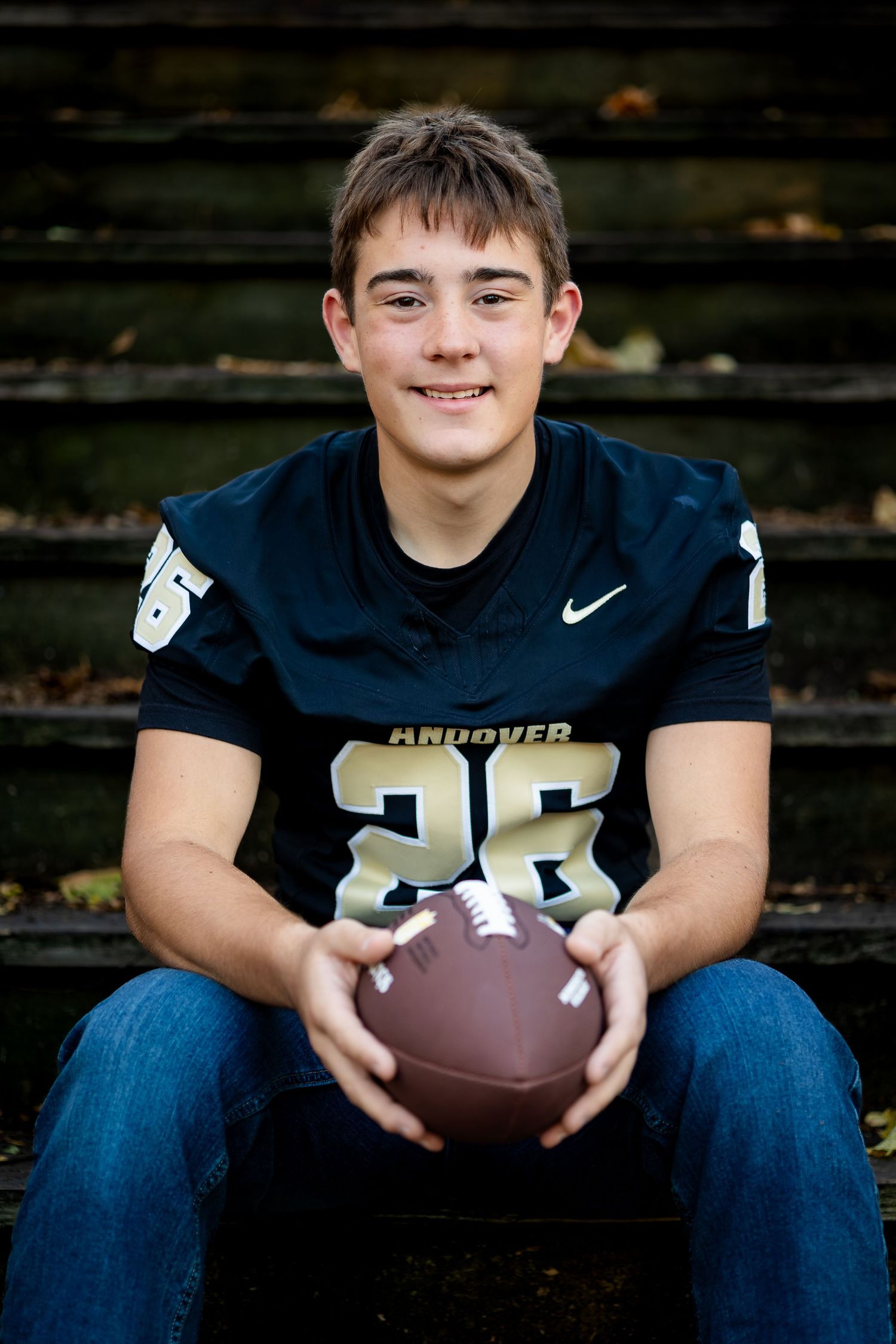 Young athlete in navy football uniform sits on bleachers holding football in outdoor sports portrait series.