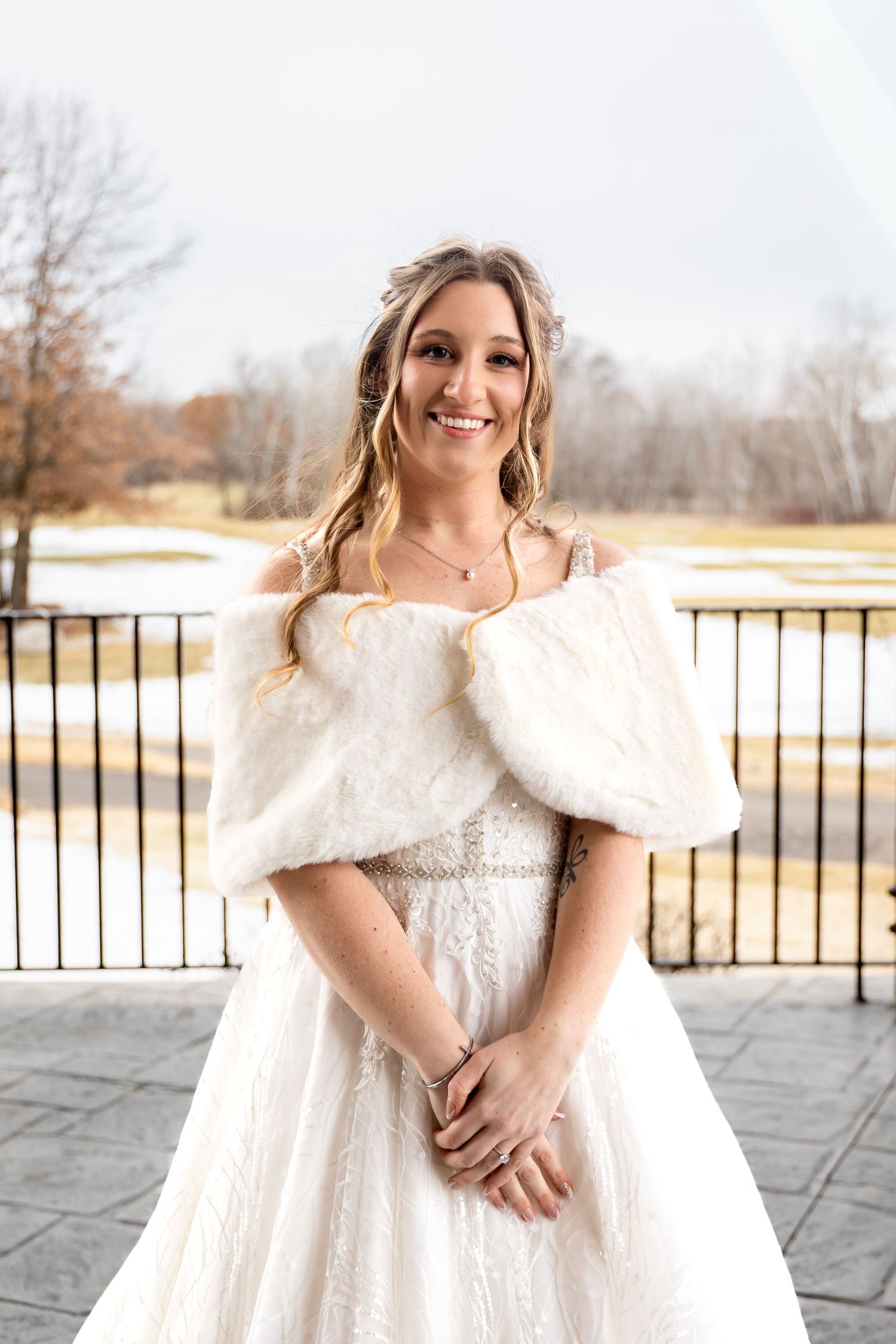 Smiling bride poses outdoors in a white lace gown with a faux fur wrap.