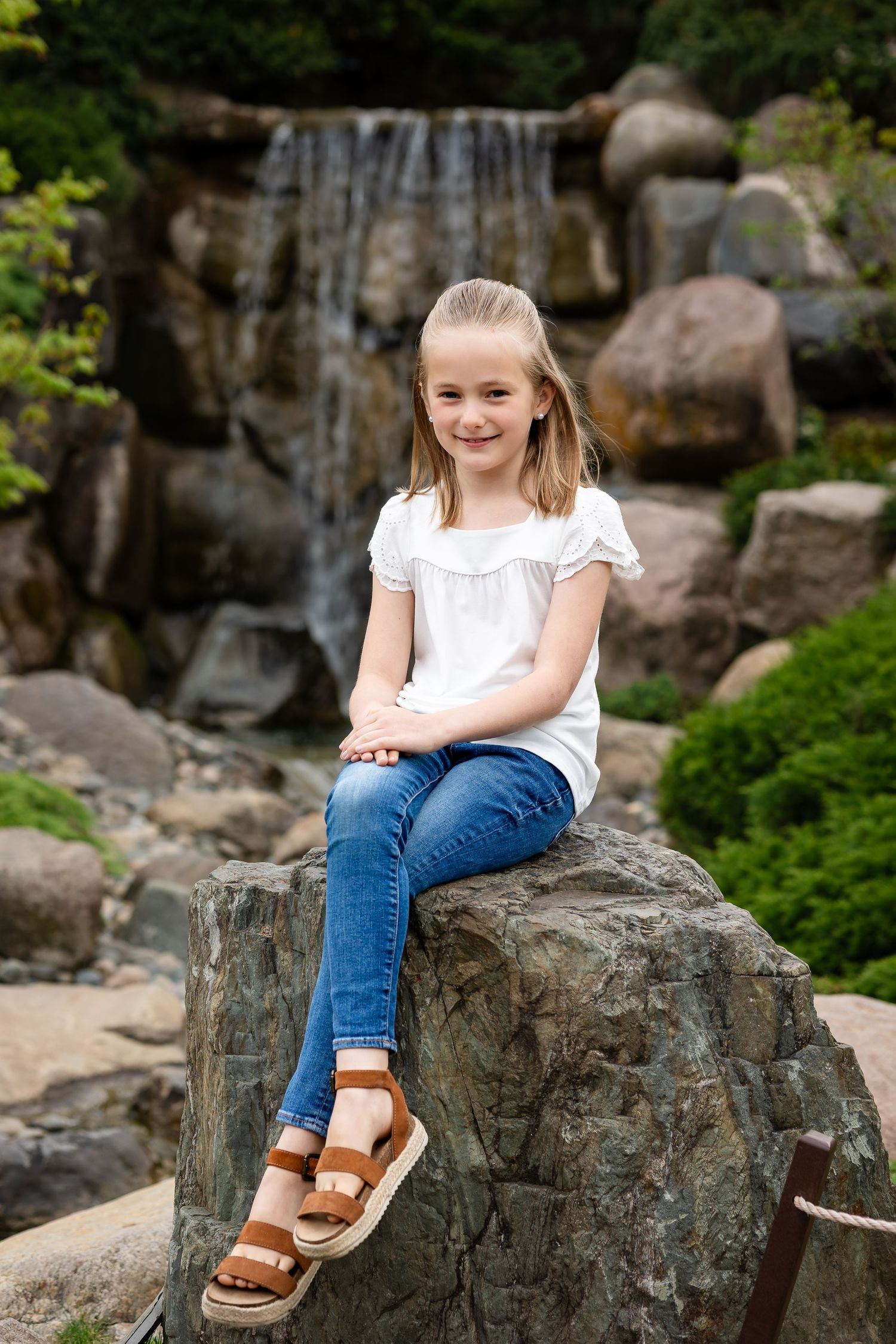 Young girl in white top and jeans sitting on a rock in front of a waterfall in a garden park.