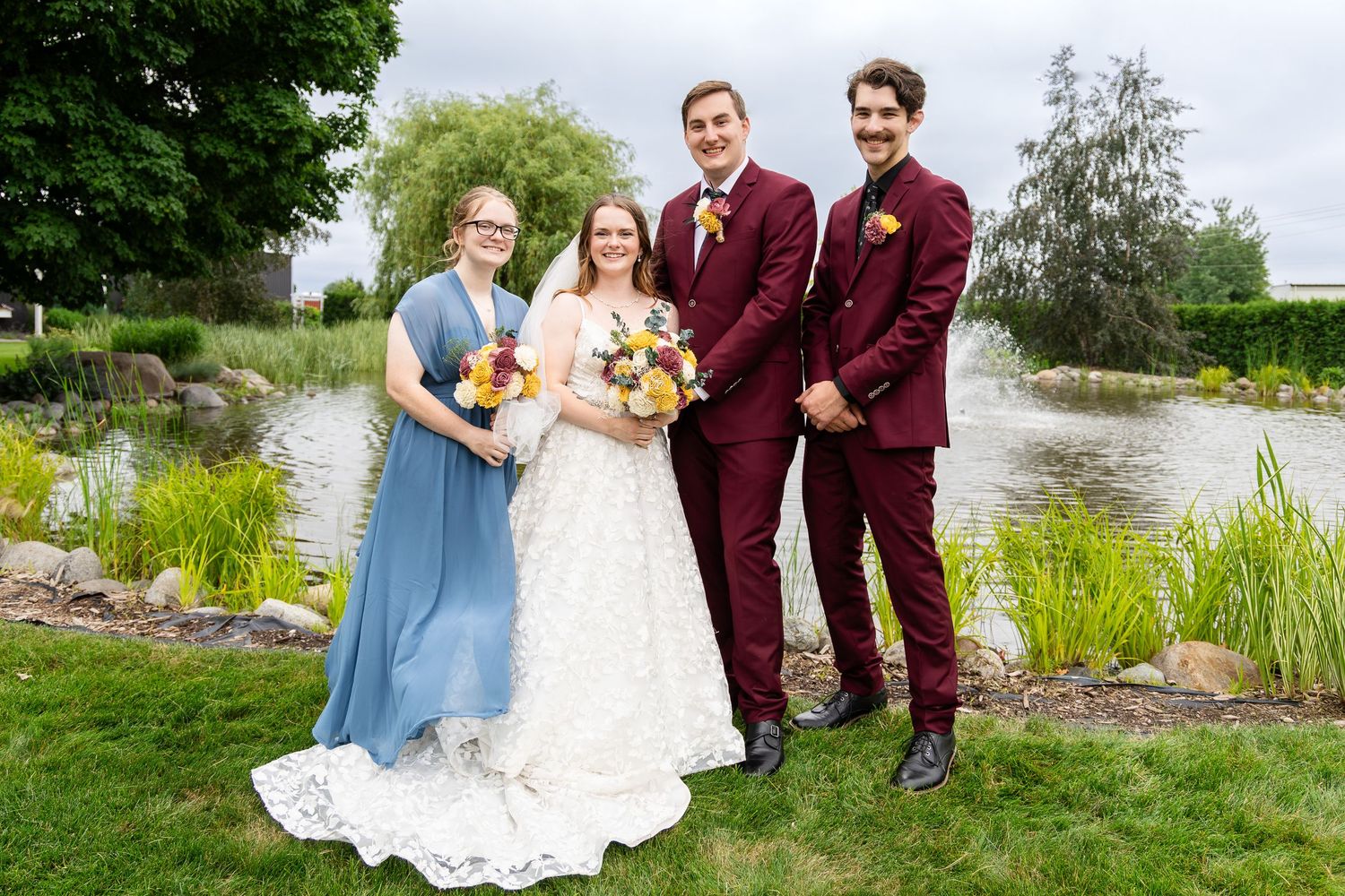 Wedding celebration on grassy lawn near pond with bridal party in burgundy and blue attire.