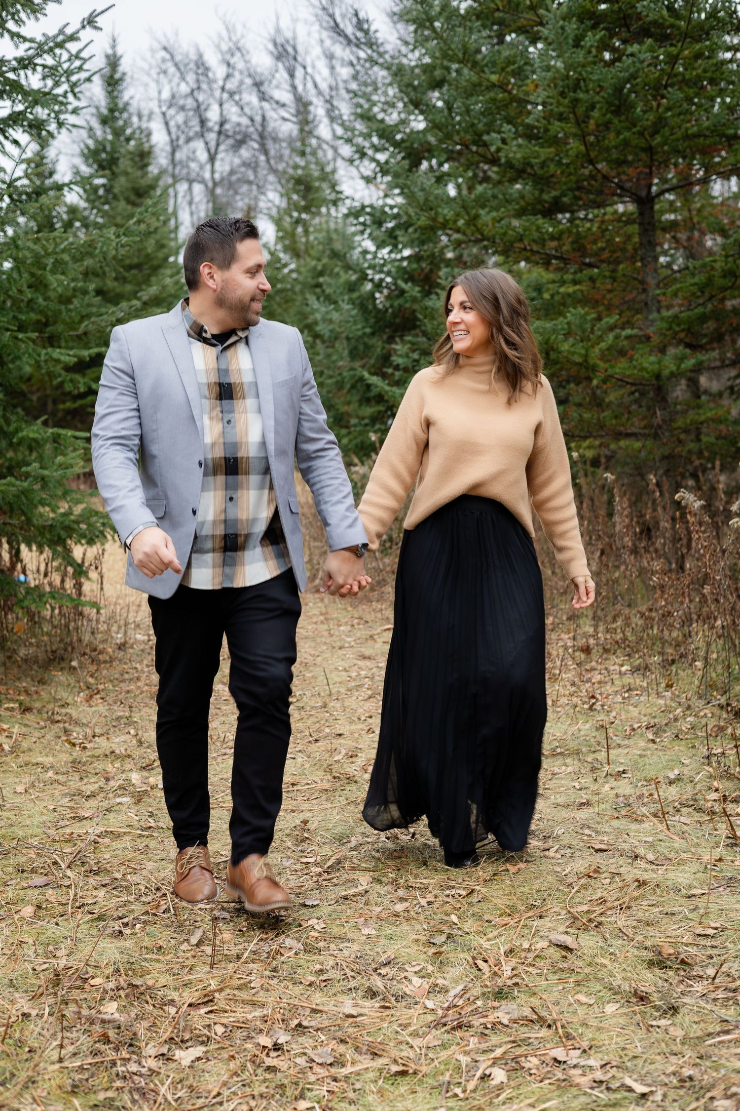 A couple holding hands while walking through a wooded area in casual attire.