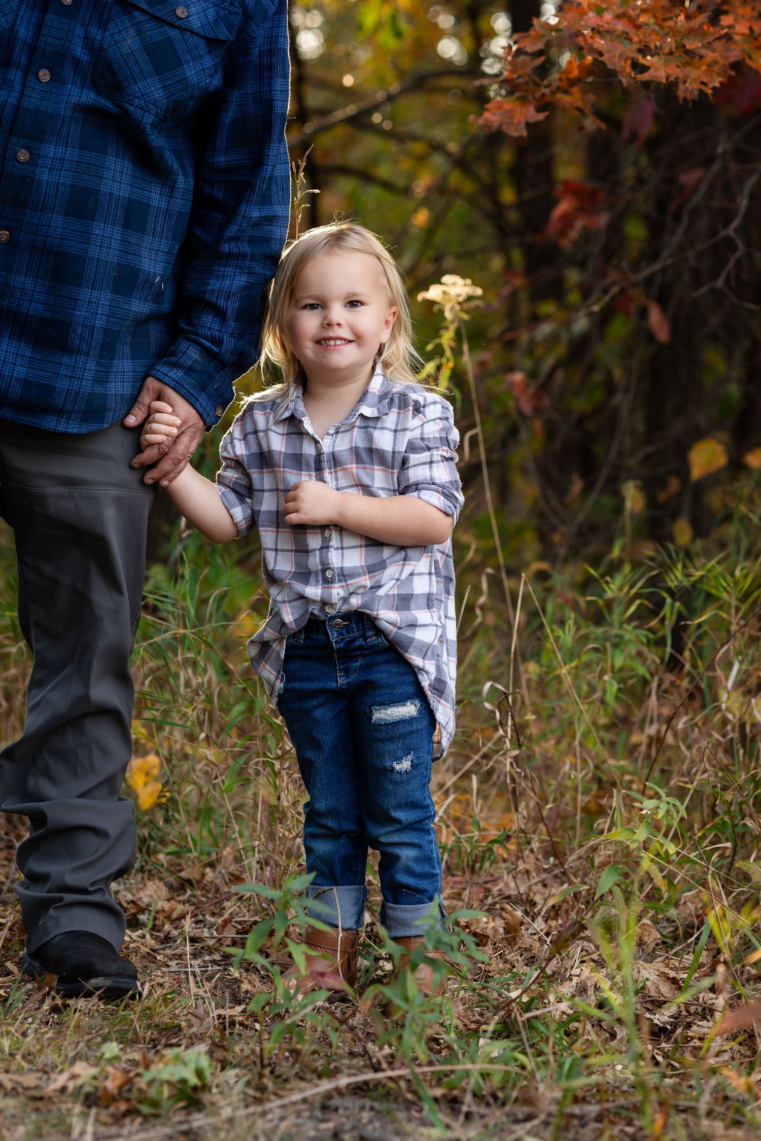 Child in plaid shirt and jeans walking through autumn foliage with adult guidance.