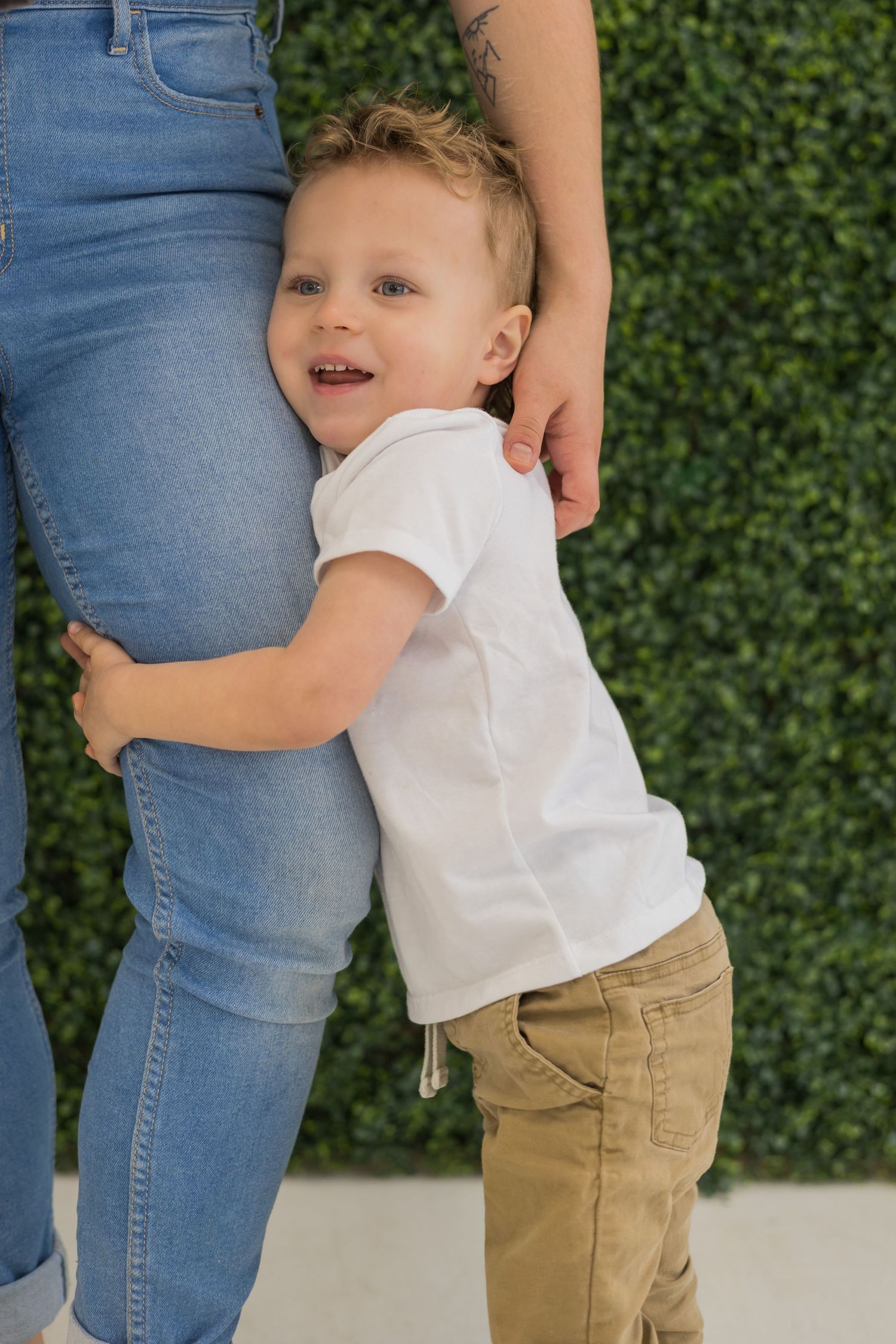 A toddler in white shirt and khaki pants hugging legs wearing blue jeans against a green backdrop.