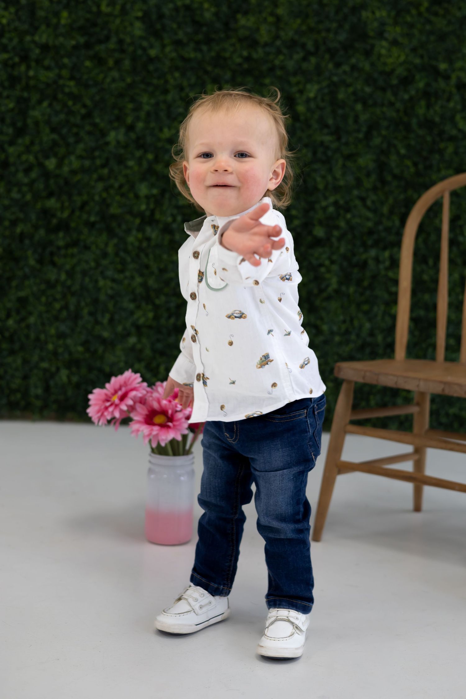 A toddler in a white patterned shirt and jeans stands confidently next to pink flowers.