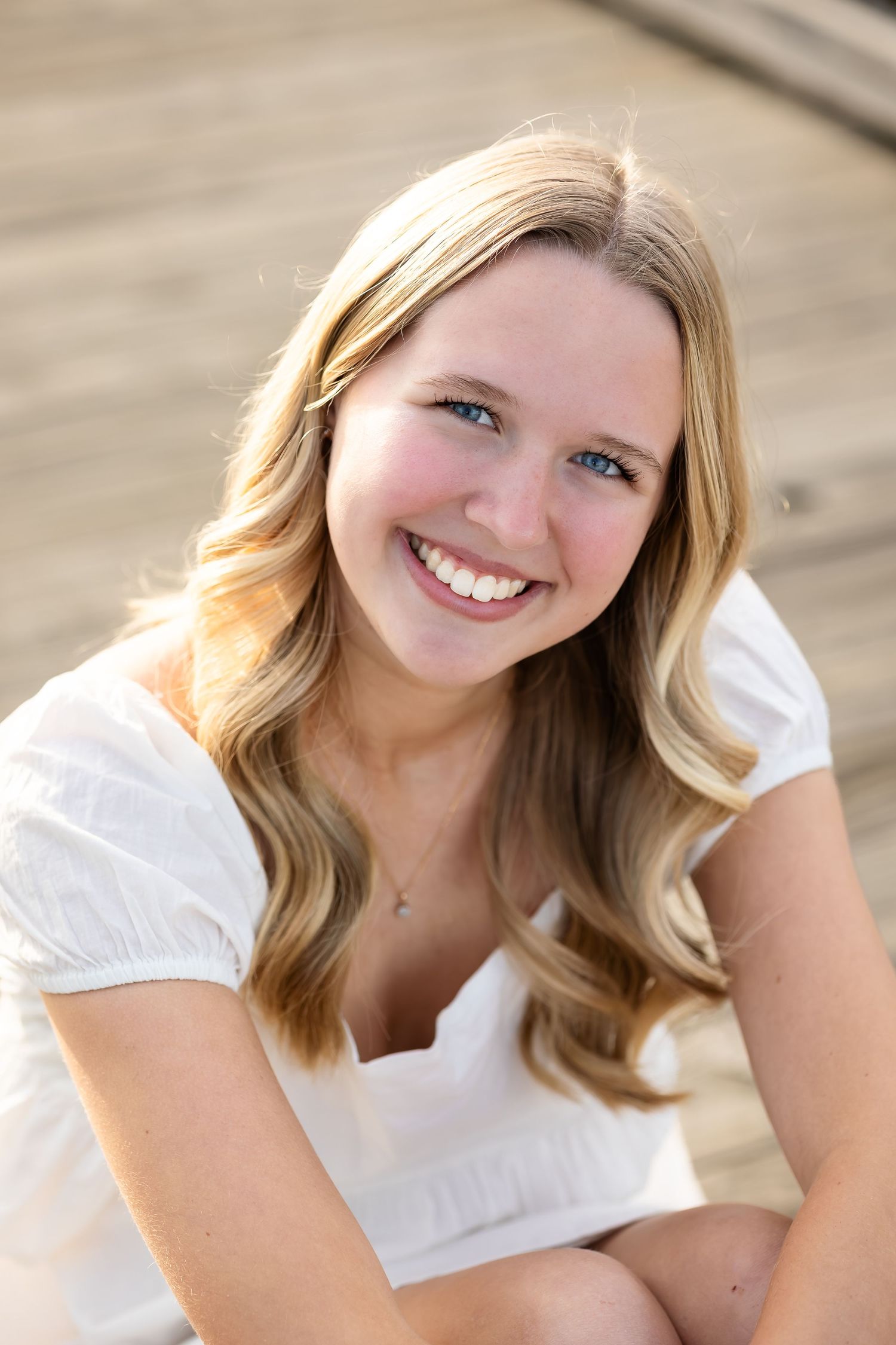A portrait of someone in a white top with wavy blonde hair smiling outdoors in warm sunlight.