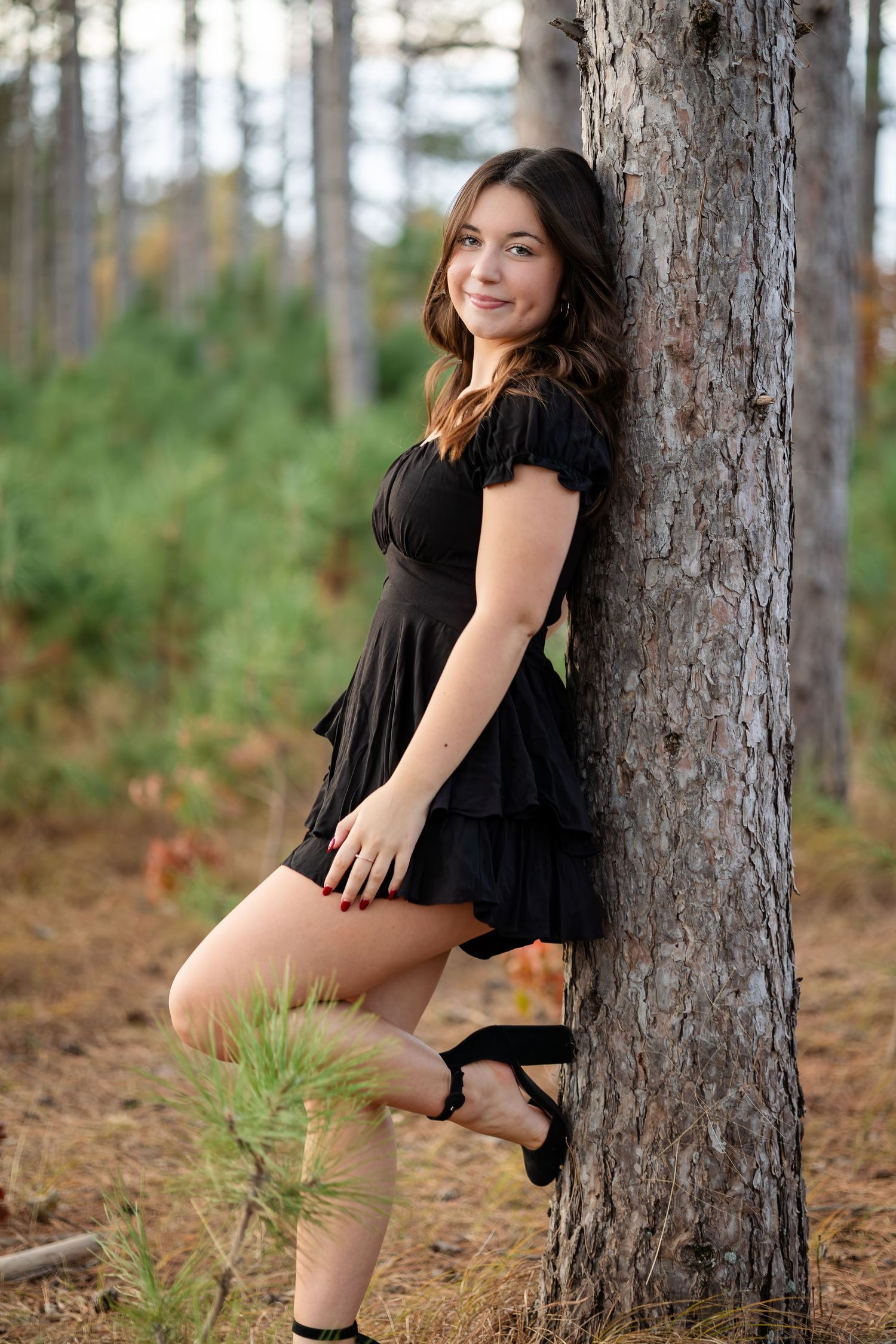Model in black dress poses against pine trees in forest setting.
