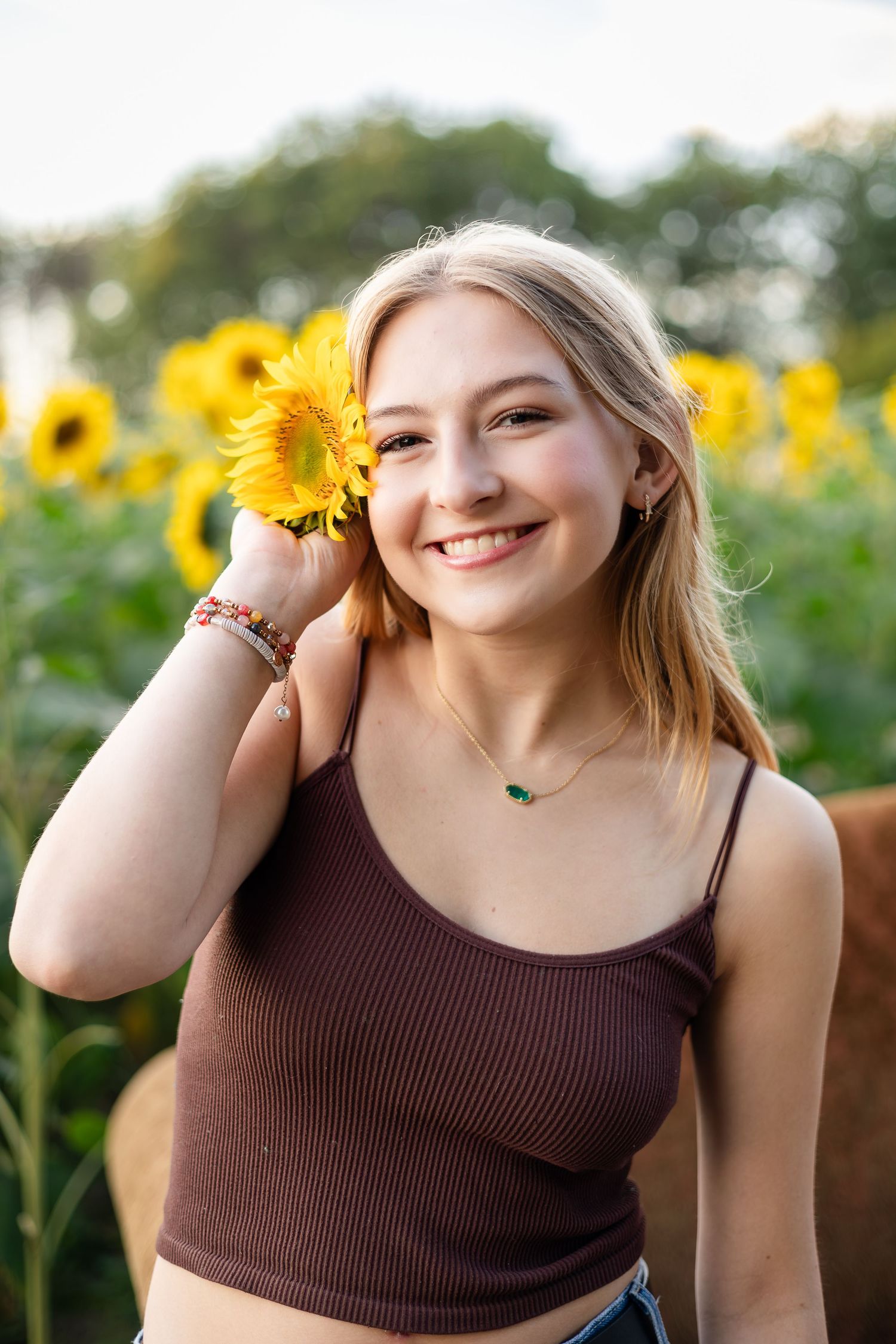 Woman in brown tank top playfully poses with sunflowers during sunset photoshoot.