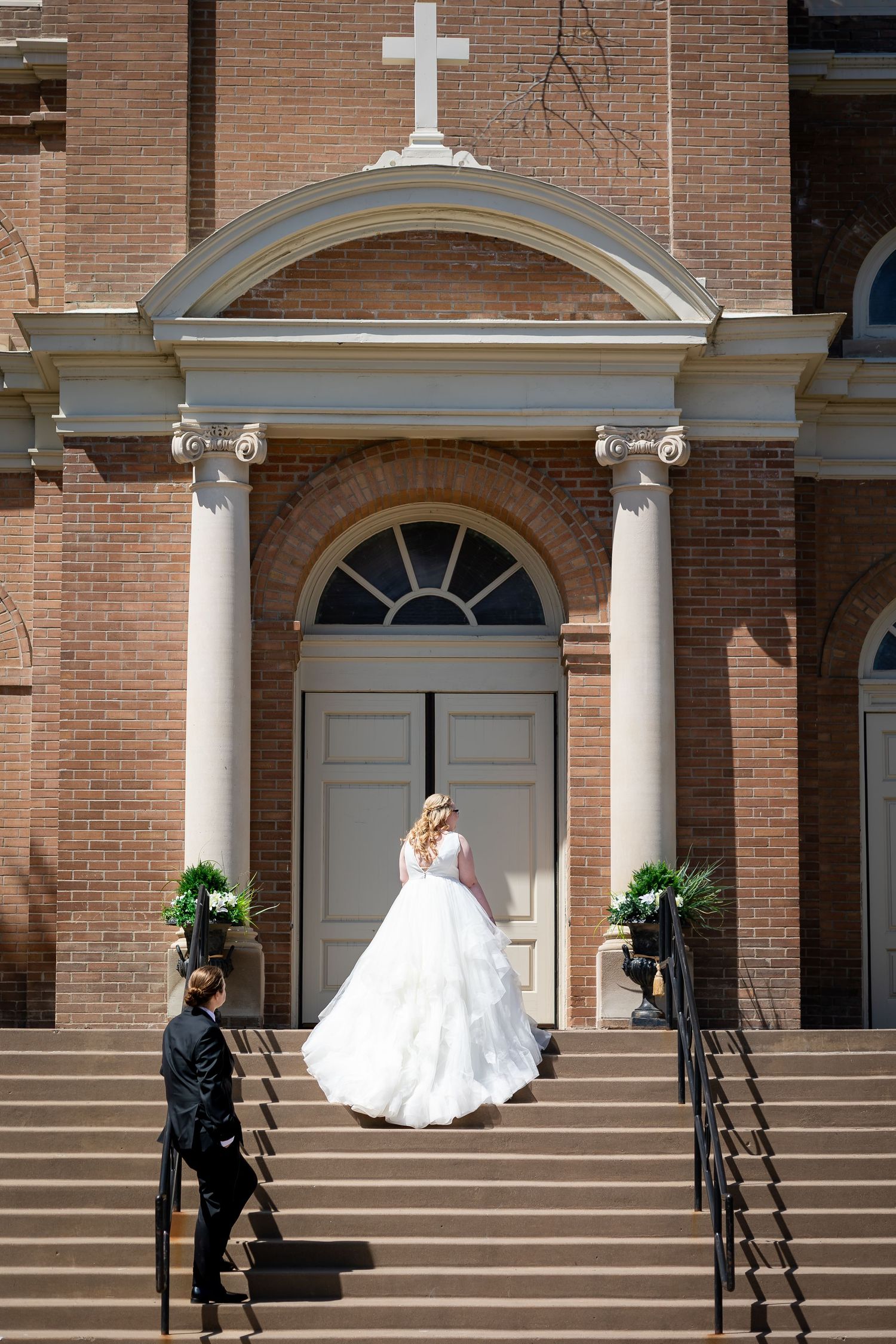 Bride in a flowing white ballgown ascending church steps with groom at a classic brick wedding venue.