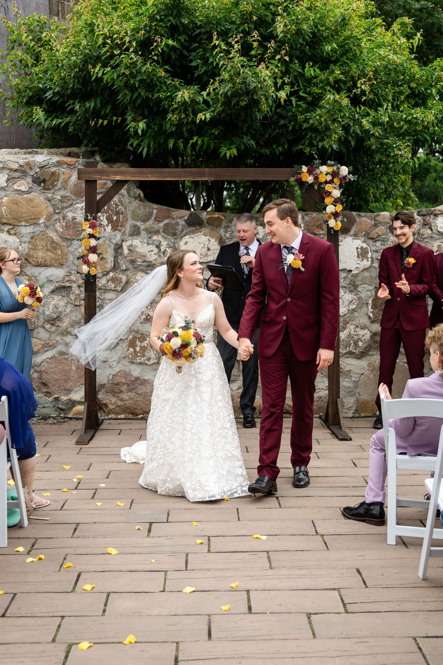 Wedding party in burgundy suits and blue dresses walks down stone pathway scattering flower petals.
