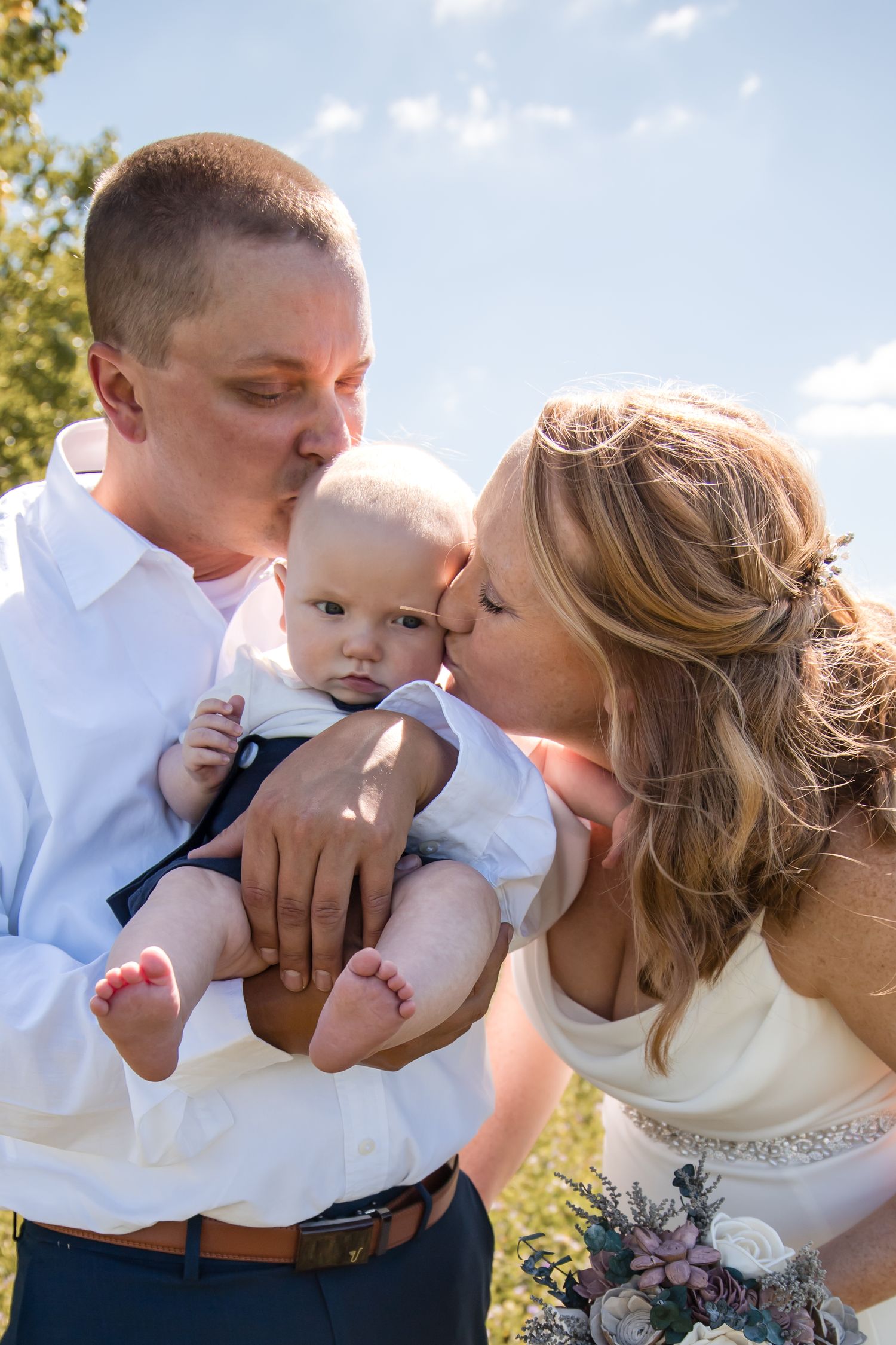 A tender wedding moment captured outdoors under blue skies with white clouds.