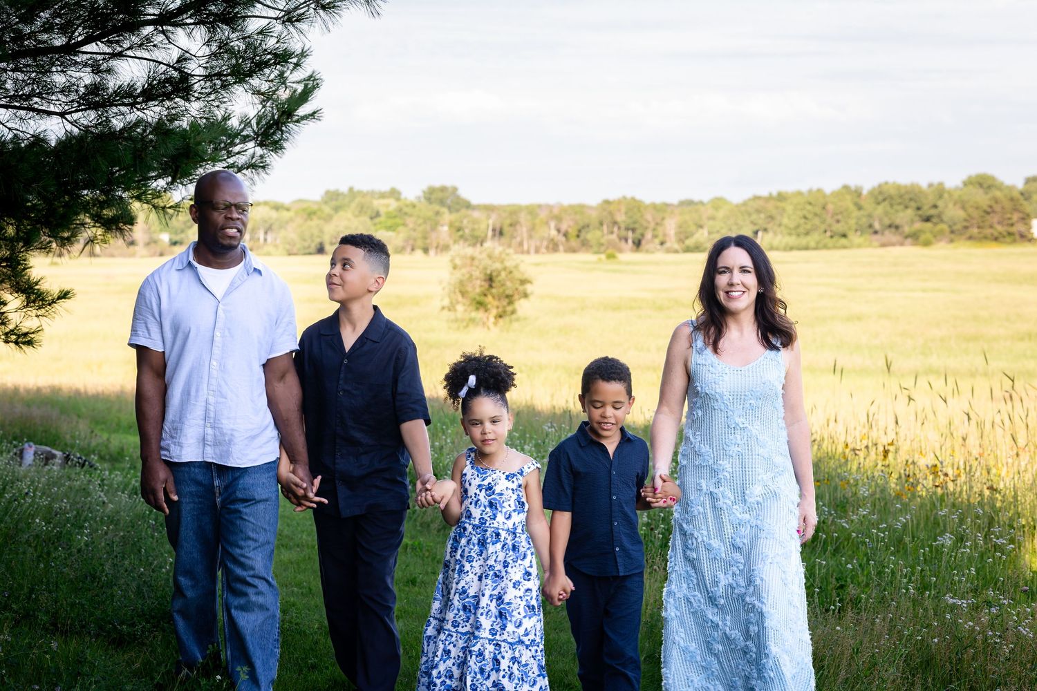 Family members walk together through a sunlit meadow wearing coordinated blue and white outfits.