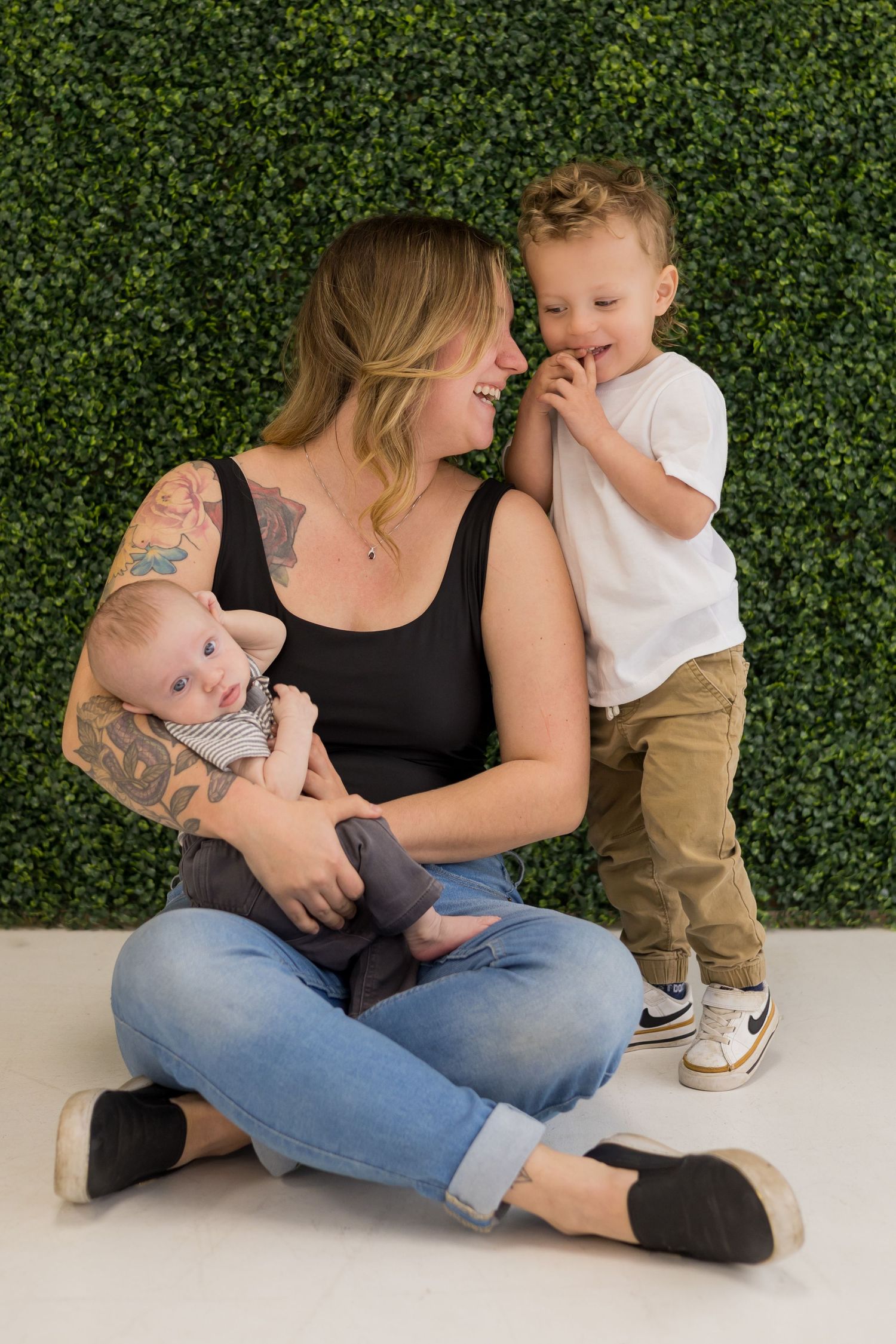 A family sitting together on the floor against a green hedge wall background.