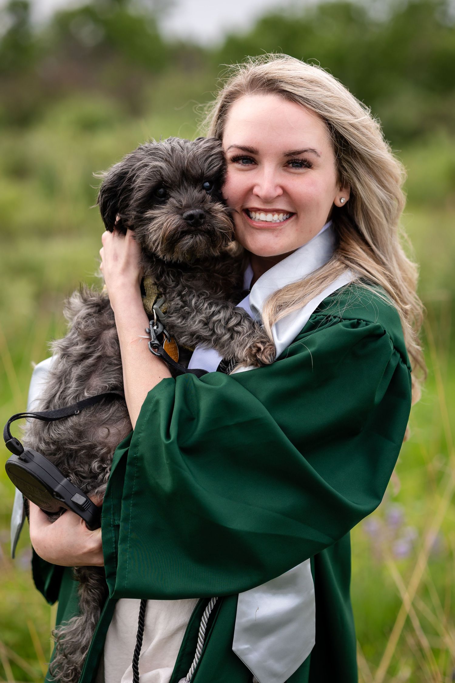 Smiling female graduate in green cap and gown holding a small gray dog outdoors.