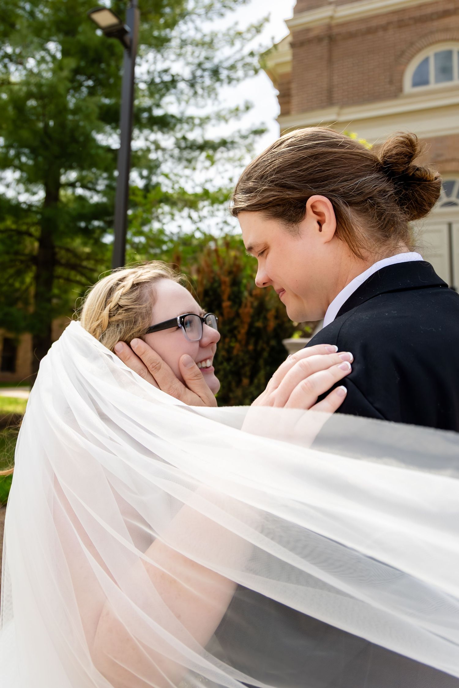 Bride and groom share a romantic moment under a flowing veil outside a brick church on their wedding day.