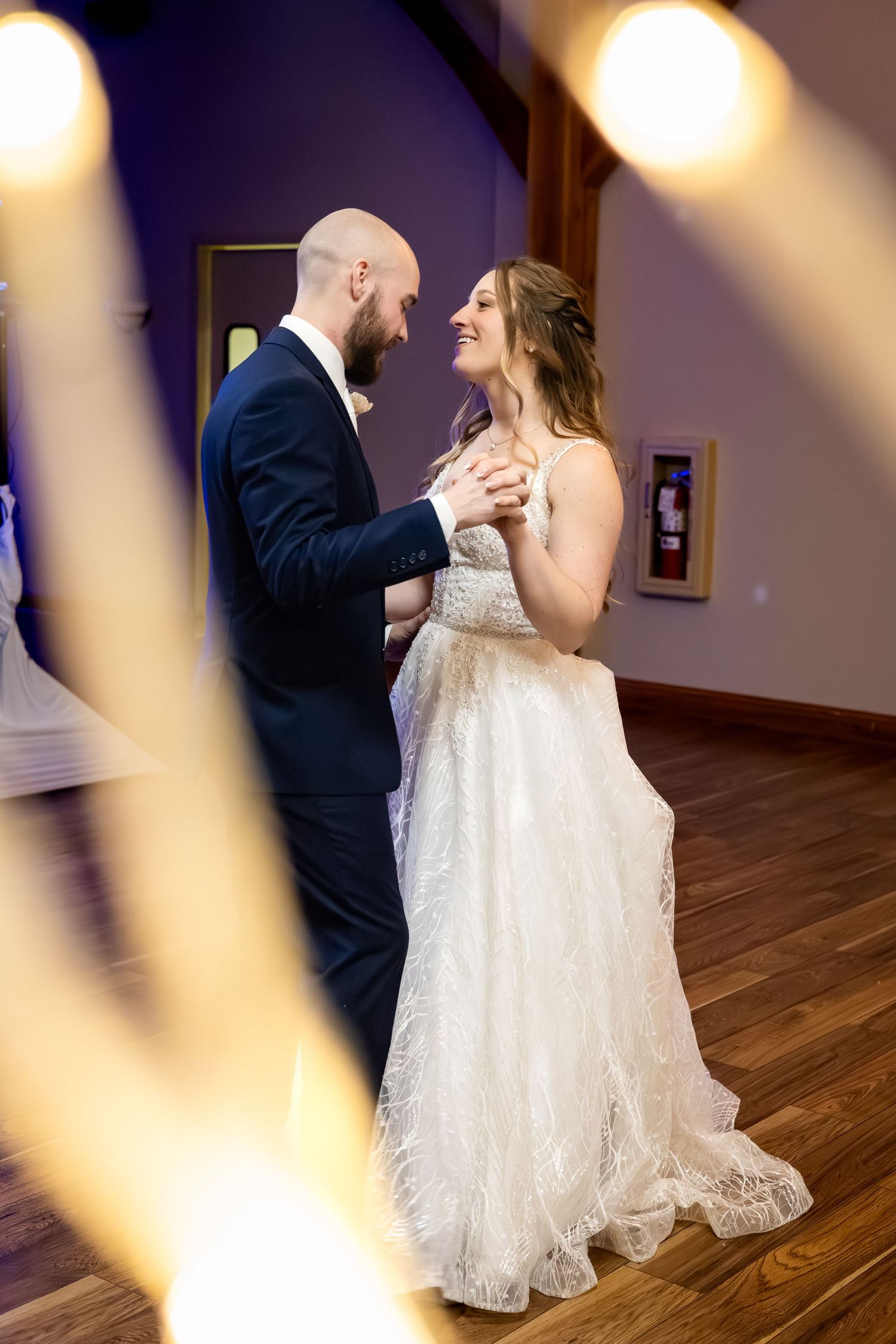 Bride in lace gown smiles at groom during first dance under warm reception lighting.
