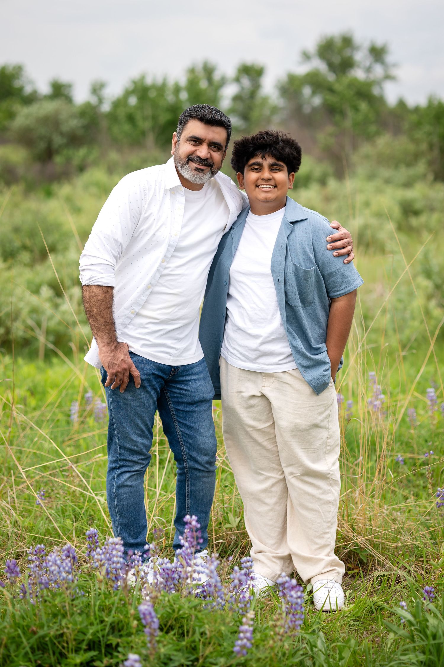 A man and teenage boy smile together in a wildflower field, the man's arm around the boy's shoulder.