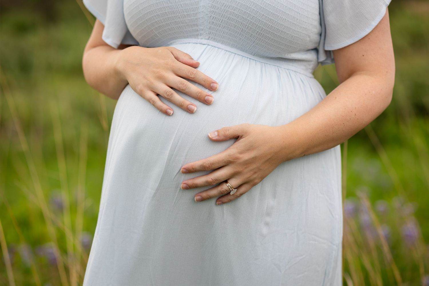 Pregnant woman in white dress cradling baby bump outdoors in green field, wearing wedding ring.