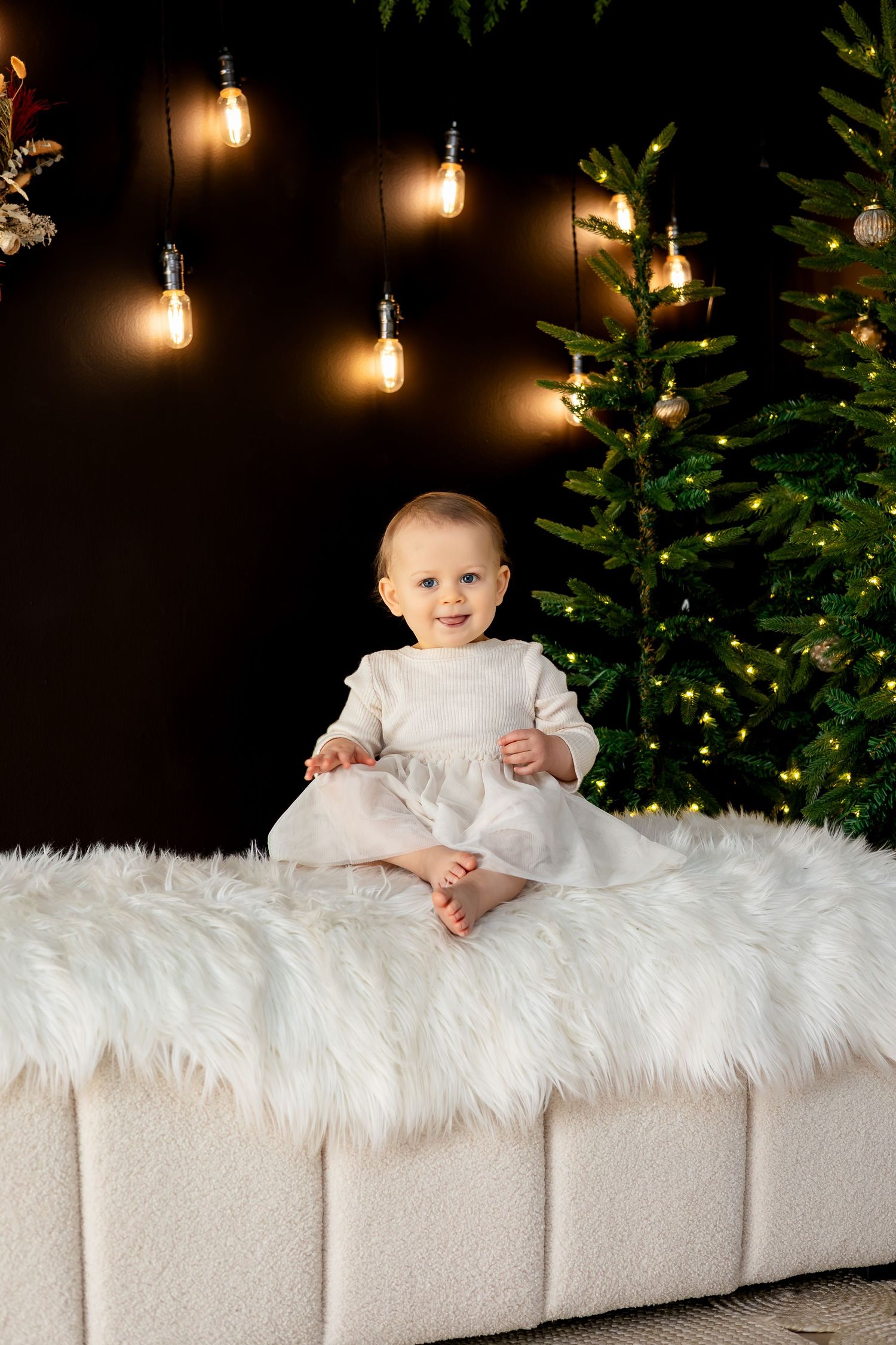 Smiling baby in white dress sitting on fluffy ottoman near decorated Christmas tree with warm glowing lights.