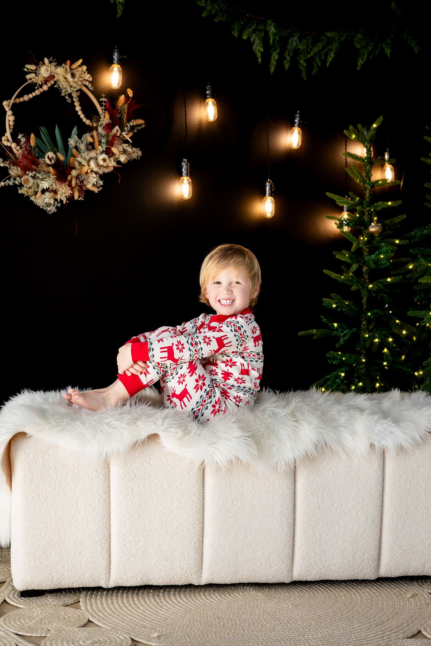Smiling toddler in Christmas pajamas sitting on a white fur bench near a decorated tree and glowing lights.