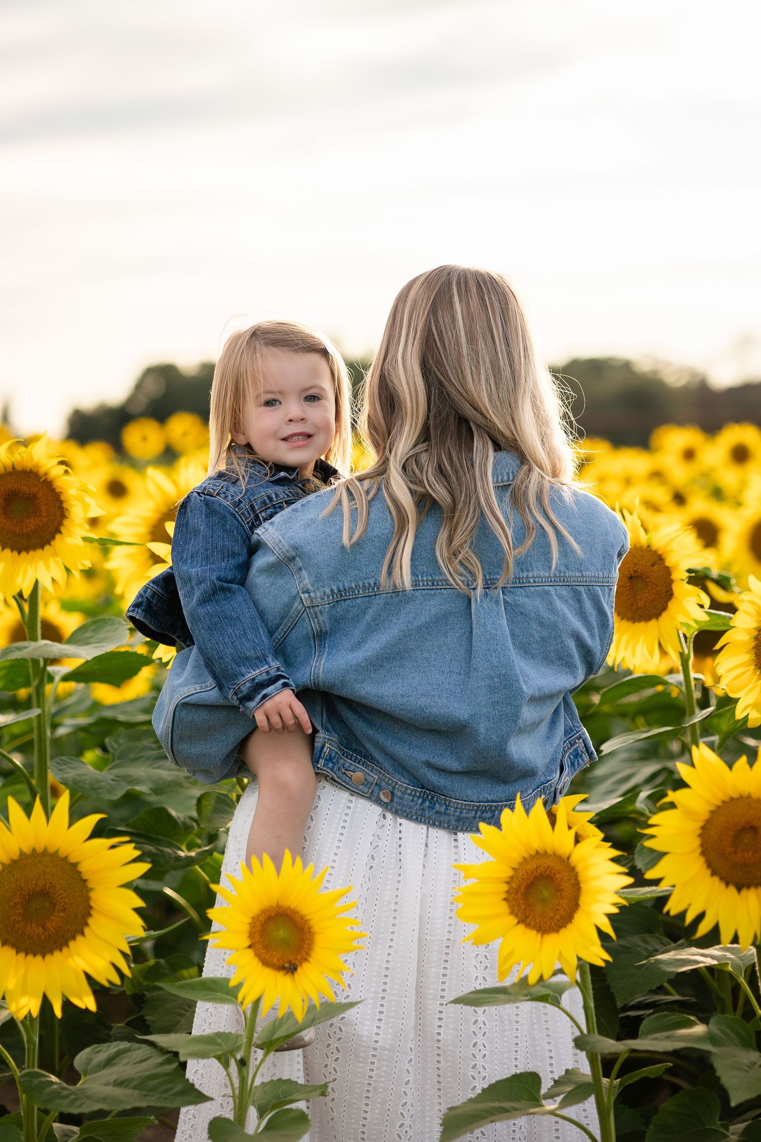 Mother and daughter in denim jackets surrounded by blooming sunflower field.