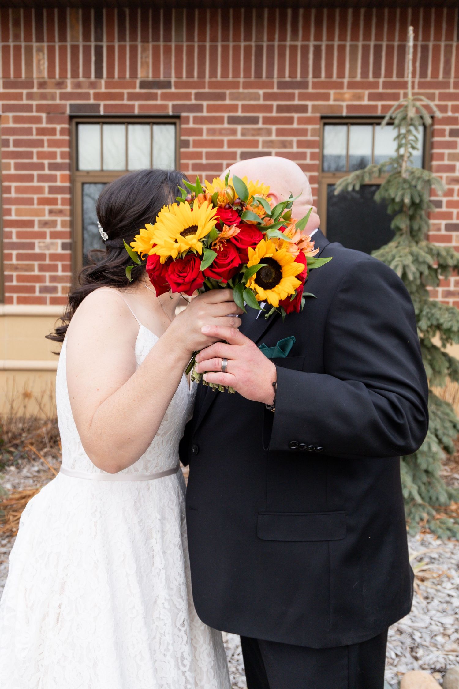 A couple shares a kiss behind a vibrant bouquet of sunflowers and red roses against brick architecture.